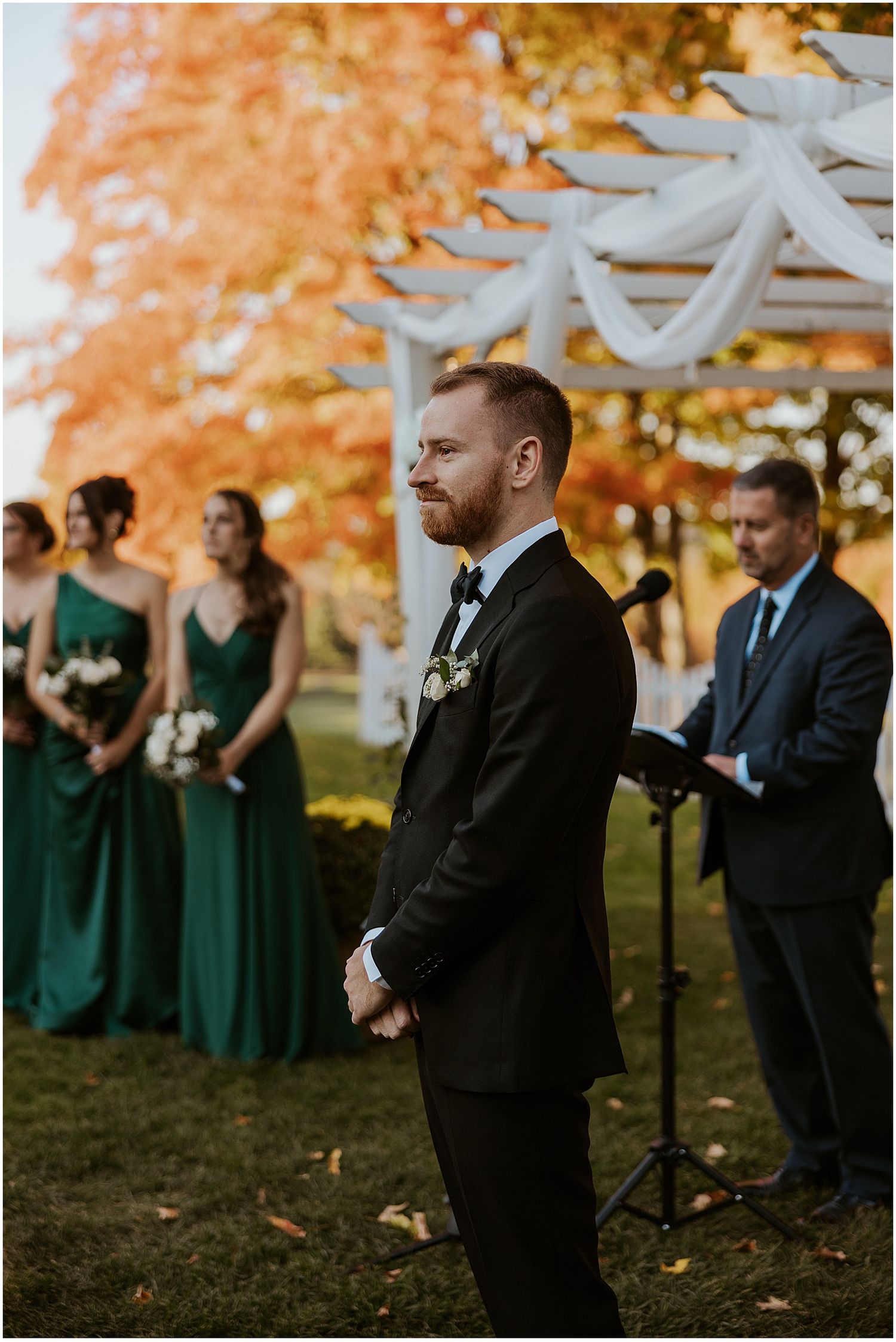 Wedding party members stand under autumn trees with bridesmaids wearing emerald green dresses.