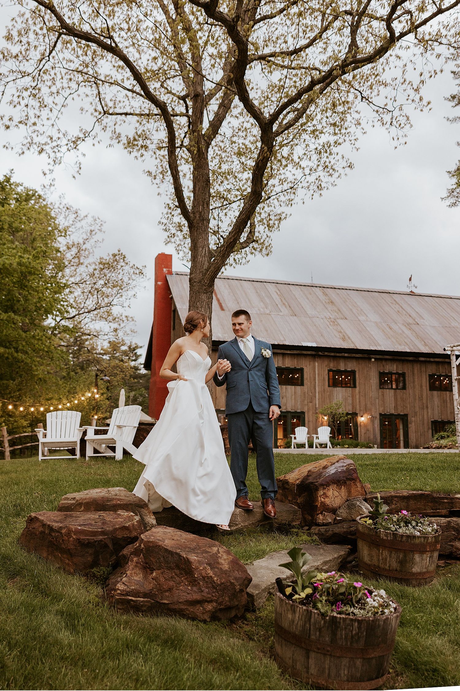 Wedding couple posing by large tree and rustic barn with decorative landscaping.