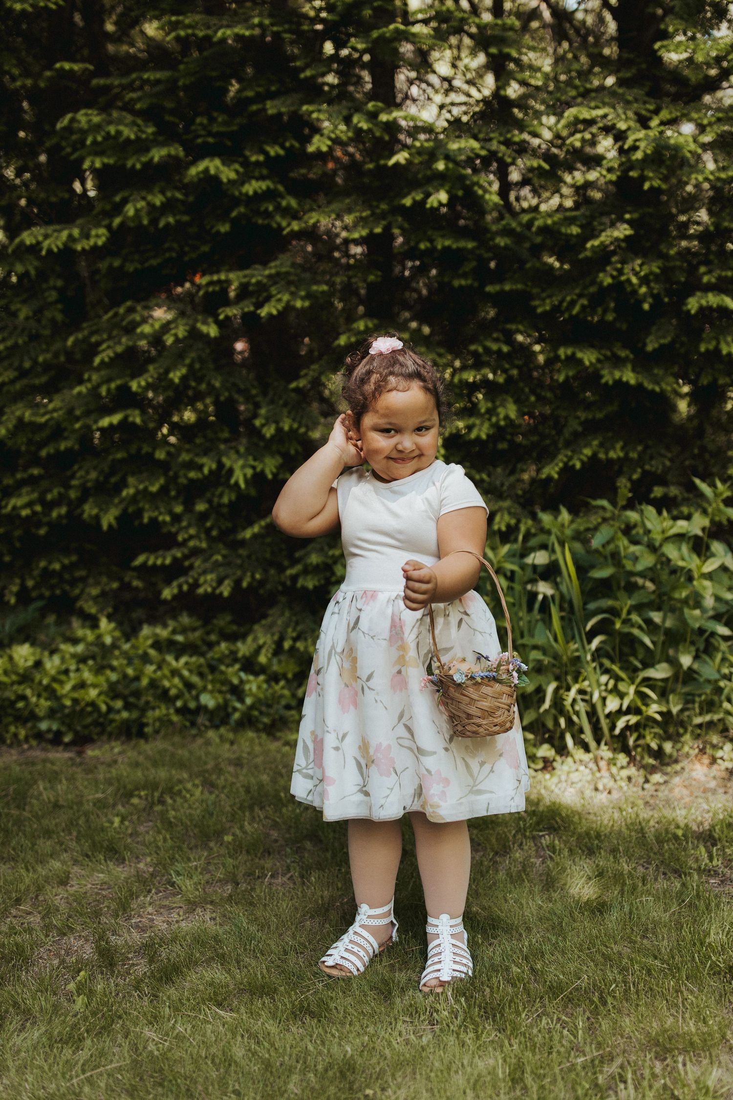 Young flower girl in white dress holding basket poses on grass at outdoor wedding.