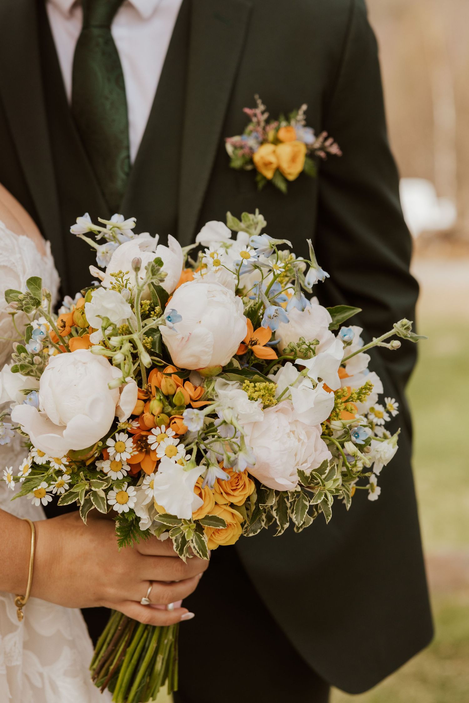 Bride holding a lush wedding bouquet with white peonies, orange roses, and daisies, beside groom in dark suit.