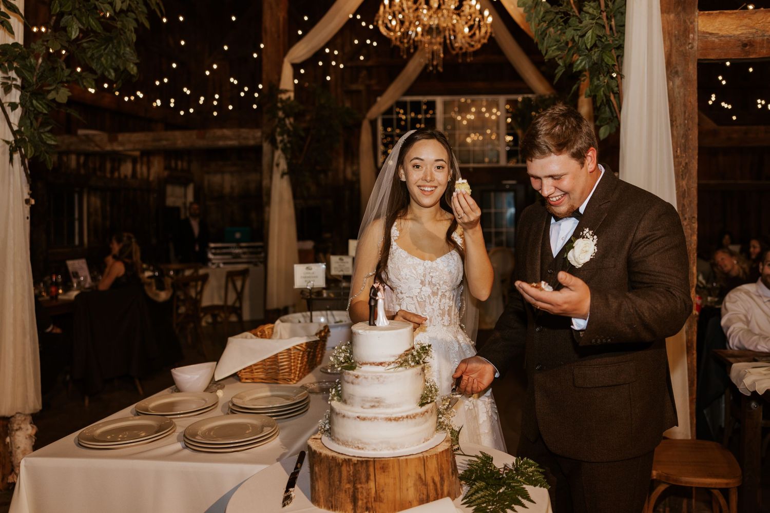 Smiling bride and groom cut a rustic tiered wedding cake in a warmly lit barn reception venue.
