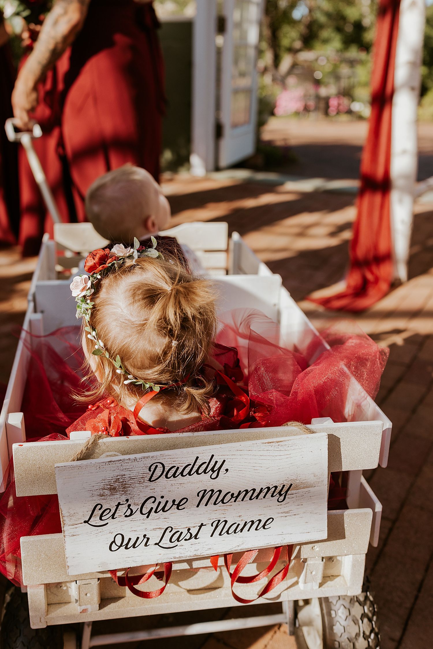 Two children in red outfits ride in a white wagon with a sign reading Daddy Let's Give Mommy Our Last Name at a NH wedding.