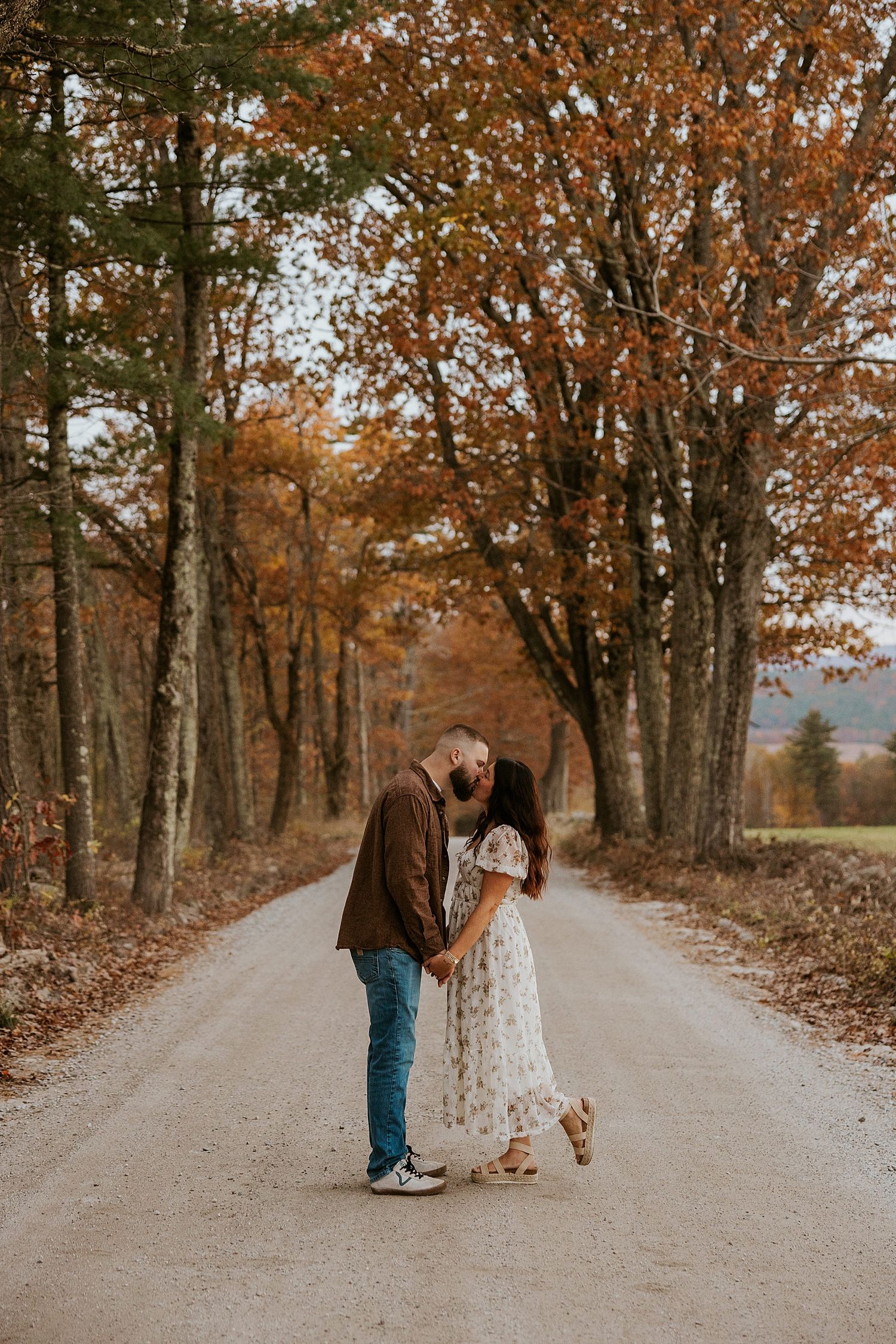 Couple kissing on a tree-lined dirt road surrounded by vibrant autumn foliage in a romantic fall engagement photo.
