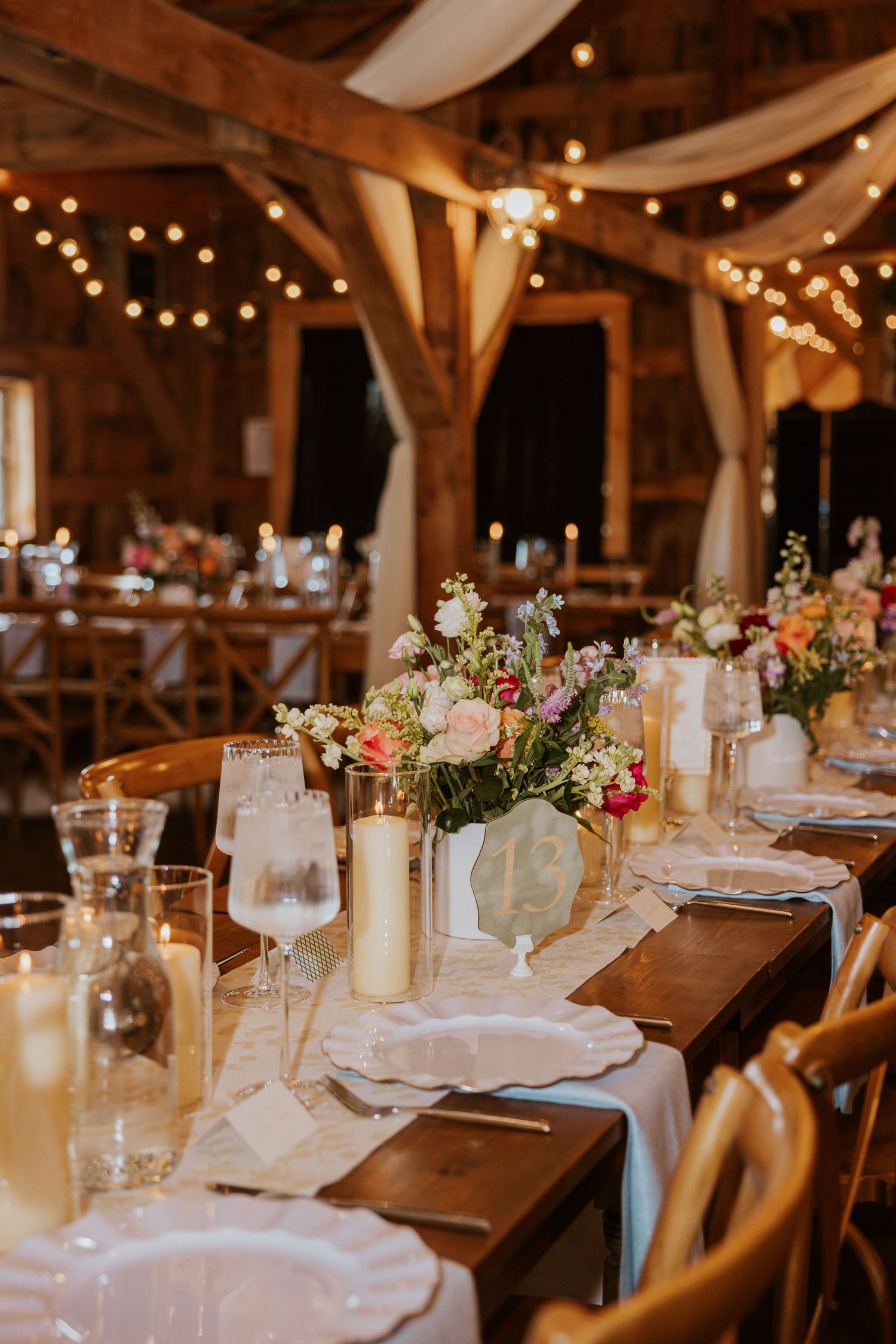 Rustic barn wedding reception with string lights, floral centerpieces, candles, and elegant table settings on wooden tables.