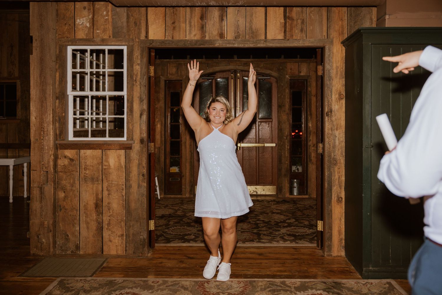 Bride in white dress and sneakers raises hands joyfully while entering rustic wooden barn wedding venue.
