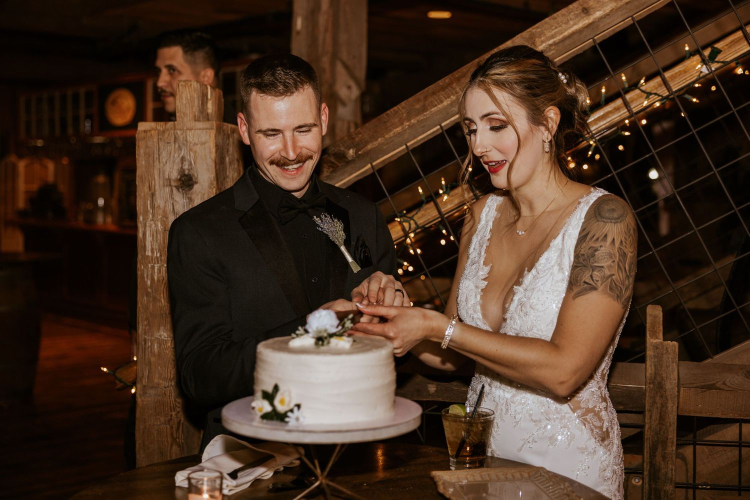 Bride and groom cutting white wedding cake together at rustic barn venue with warm string lights.