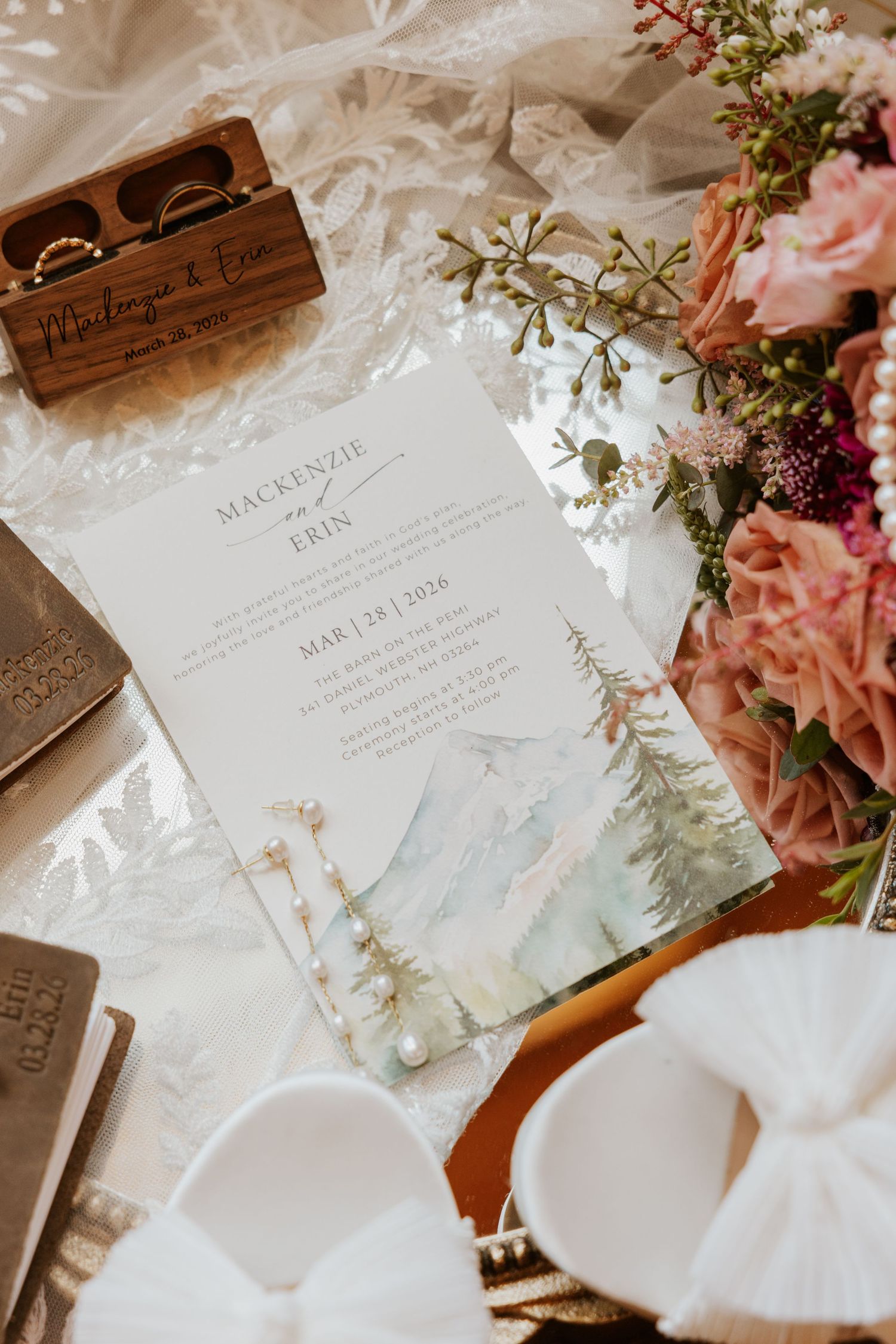 Wedding invitation with watercolor mountain design surrounded by pink flowers and wooden decor details.