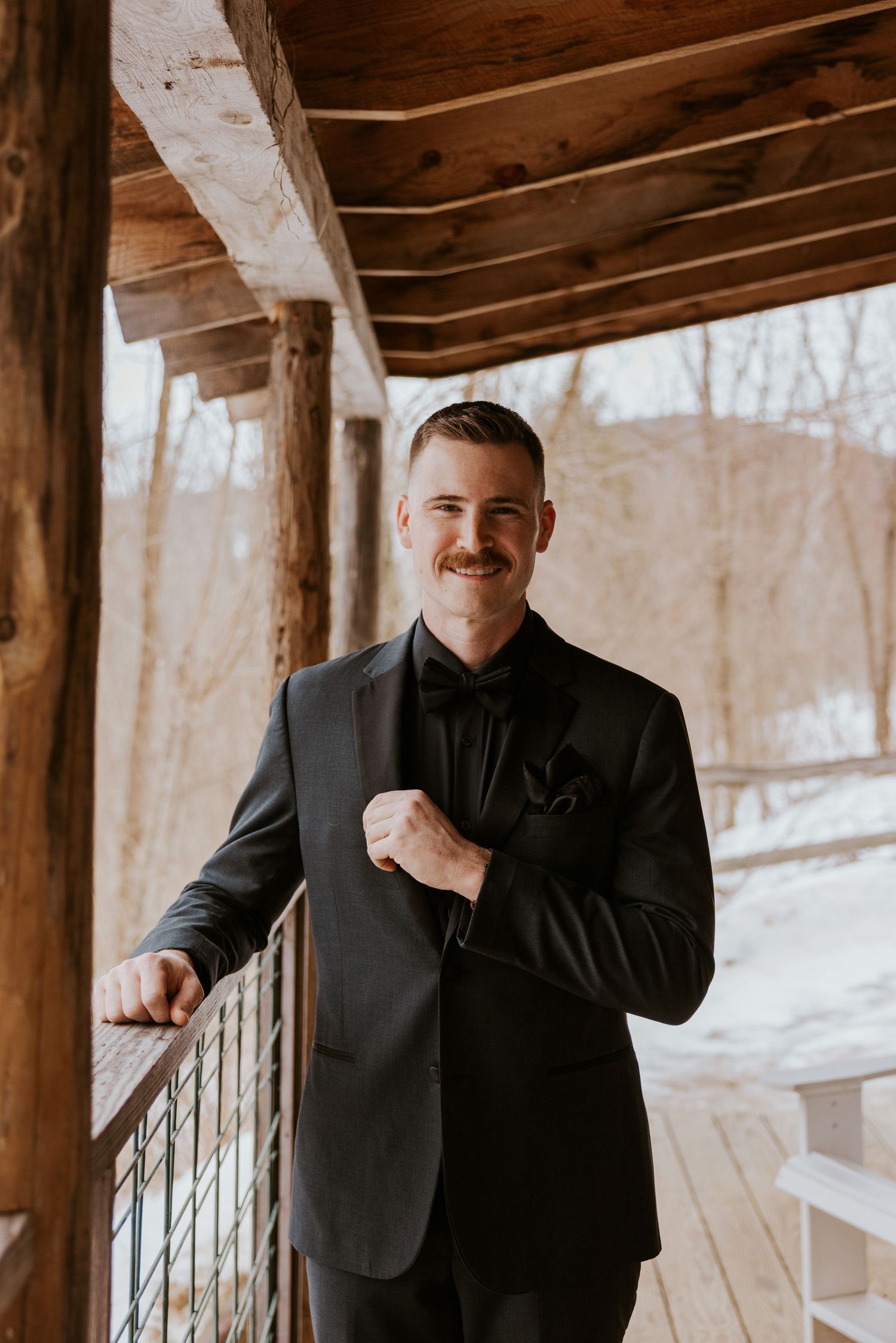 Dapper groom with mustache in black tuxedo and bow tie posing on a rustic wooden porch in winter.