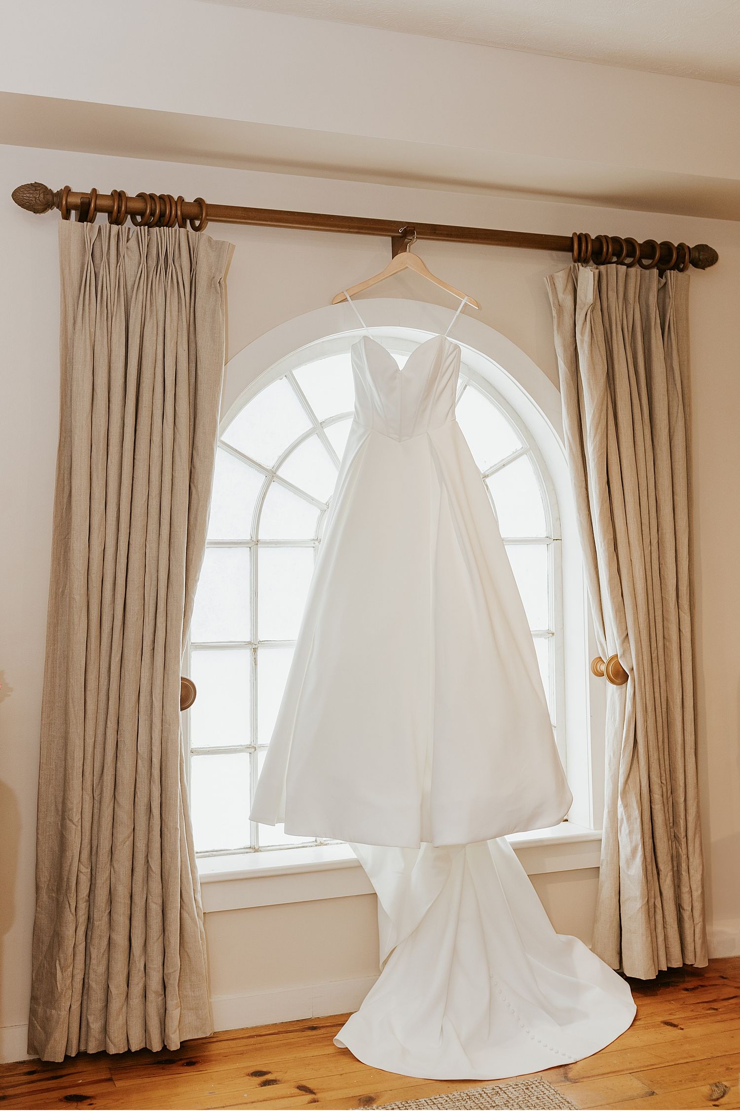 Elegant white wedding dress hanging in a sunlit room with arched windows and beige curtains.