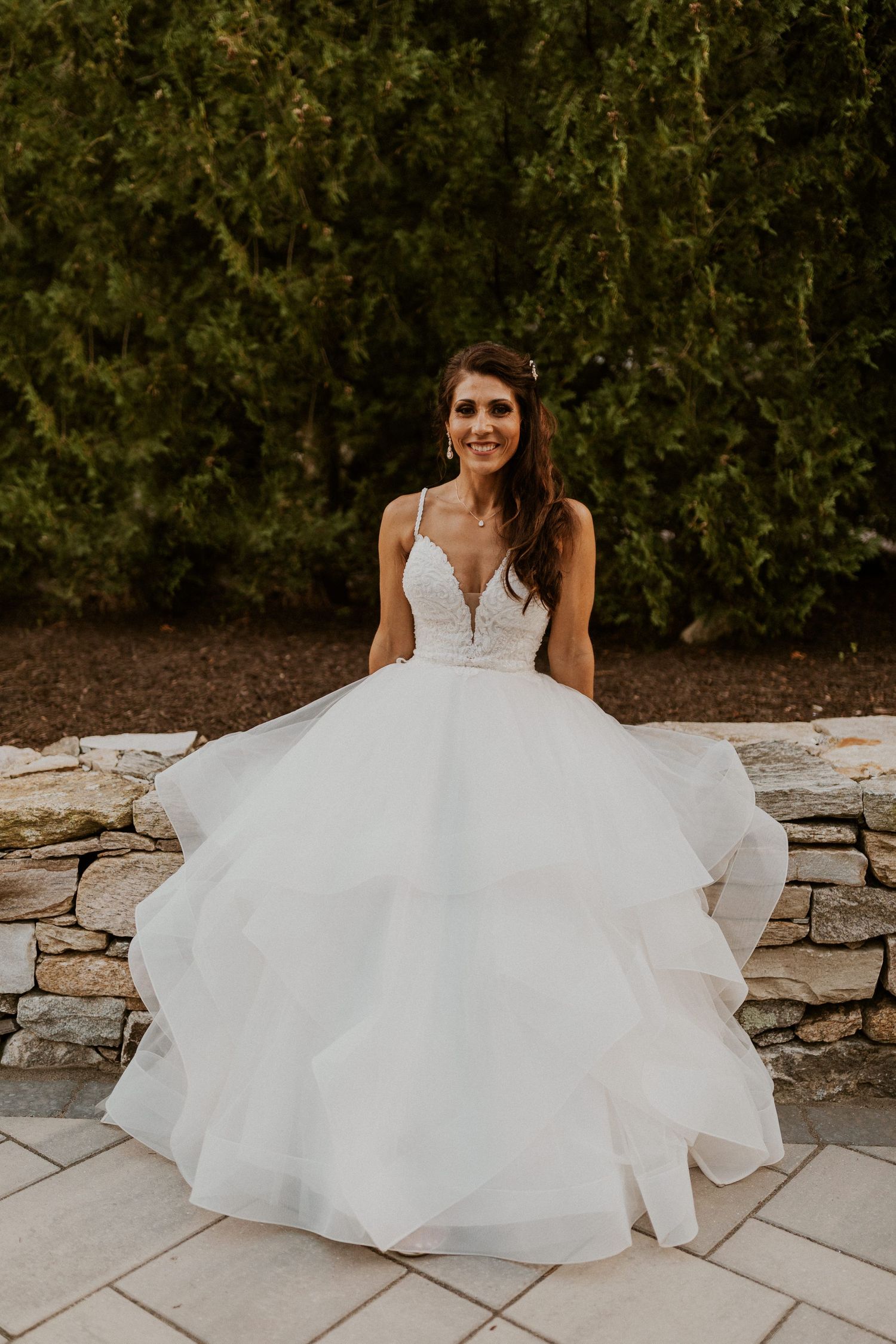 Bride in flowing white dress sits elegantly on stone wall during outdoor wedding photoshoot.