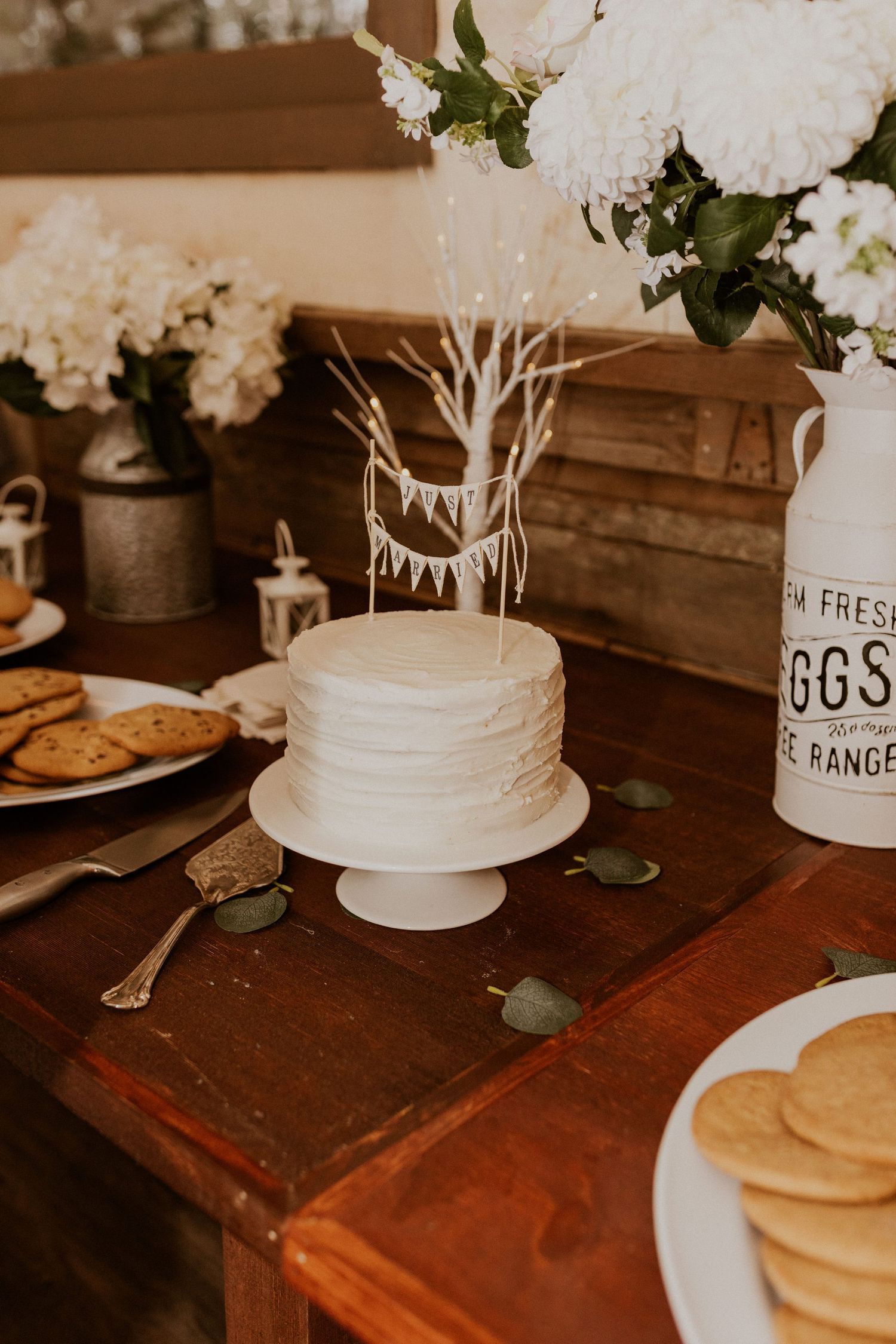 Elegant white wedding cake and rustic table decor with flowers and vintage containers at a reception.