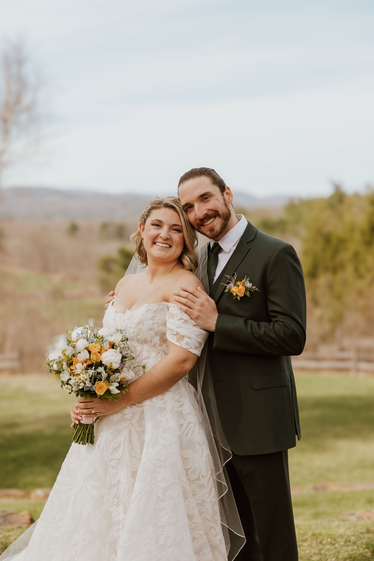 Bride in lace off-shoulder gown holds yellow and white bouquet with groom in dark suit outdoors.