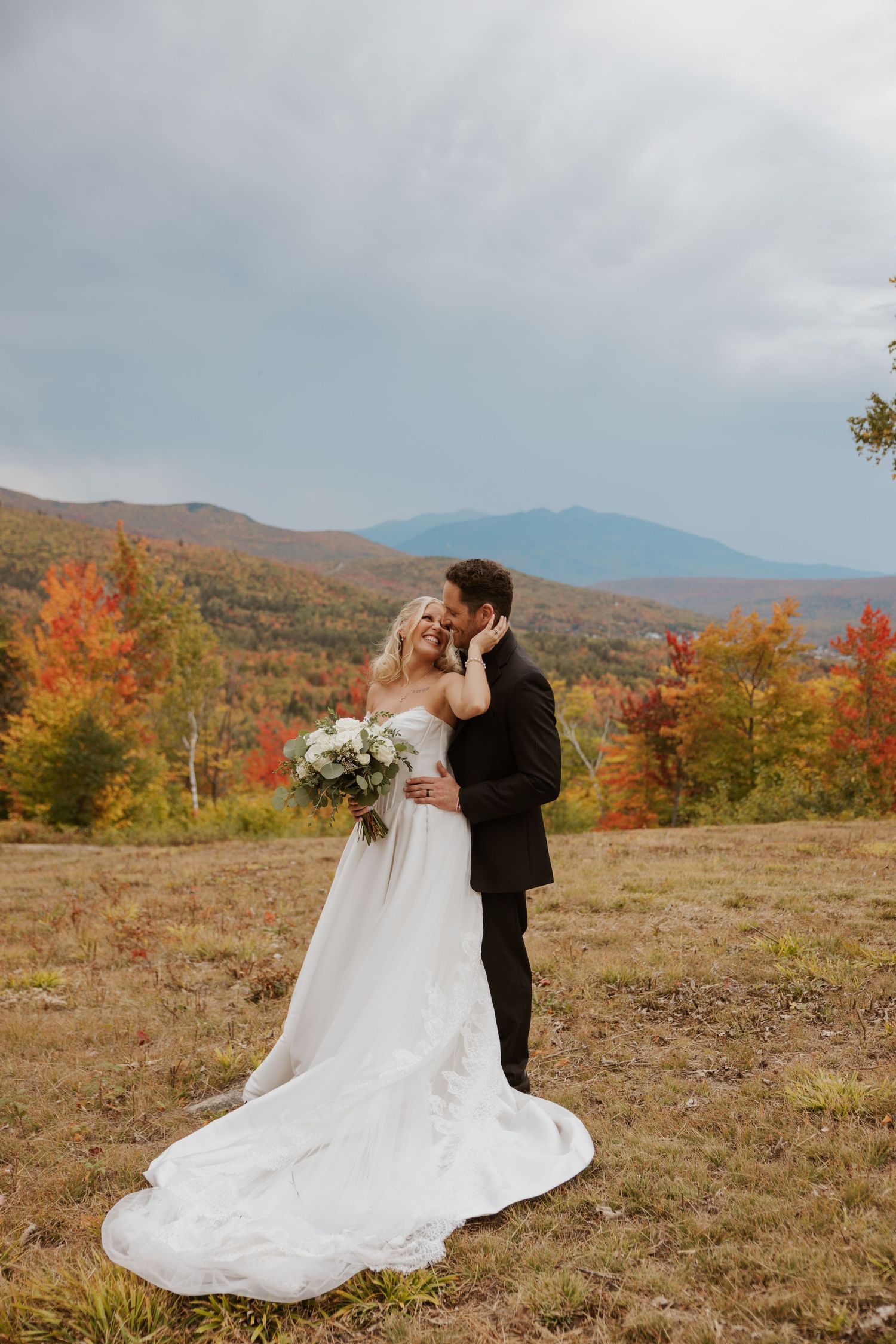 Bride and groom embracing on a hillside with vibrant autumn foliage and mountain backdrop on their wedding day.