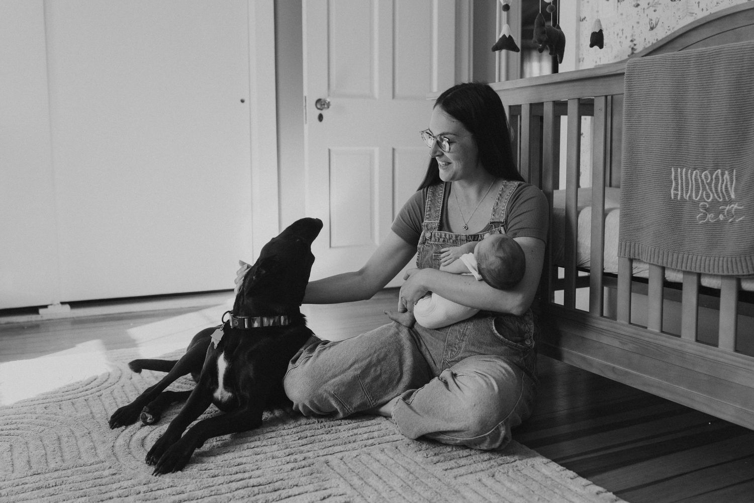 Black and white photo of mother breastfeeding newborn on floor beside black dog near wooden crib.