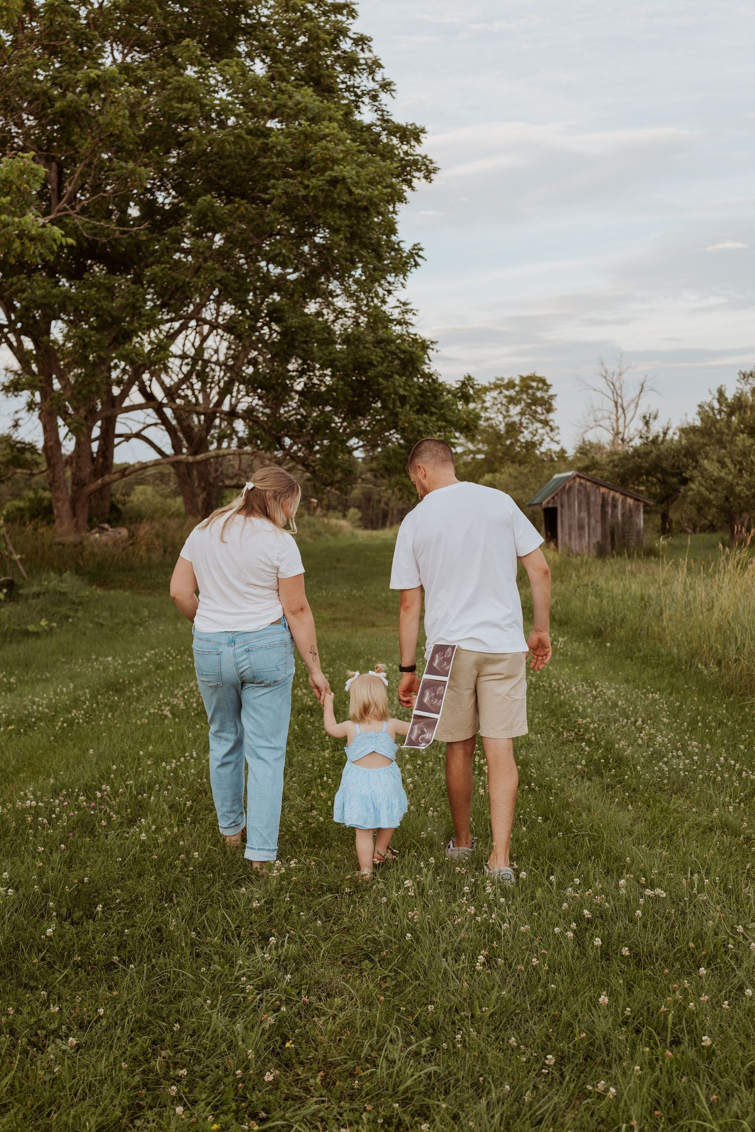 Family of three walks through green meadow, toddler holding ultrasound photos between parents.