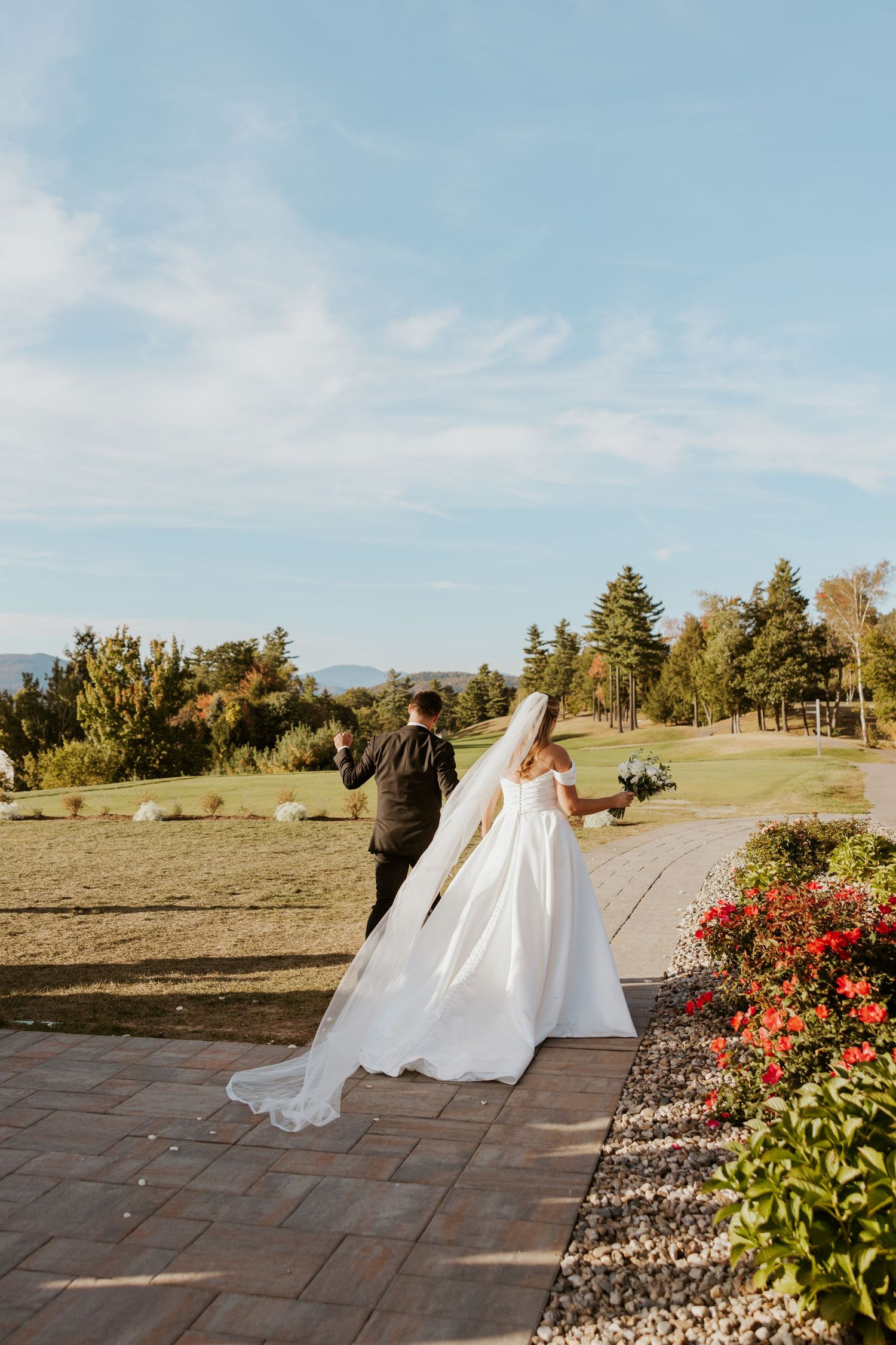 Bride and groom walk hand in hand along garden path with scenic mountain landscape backdrop.