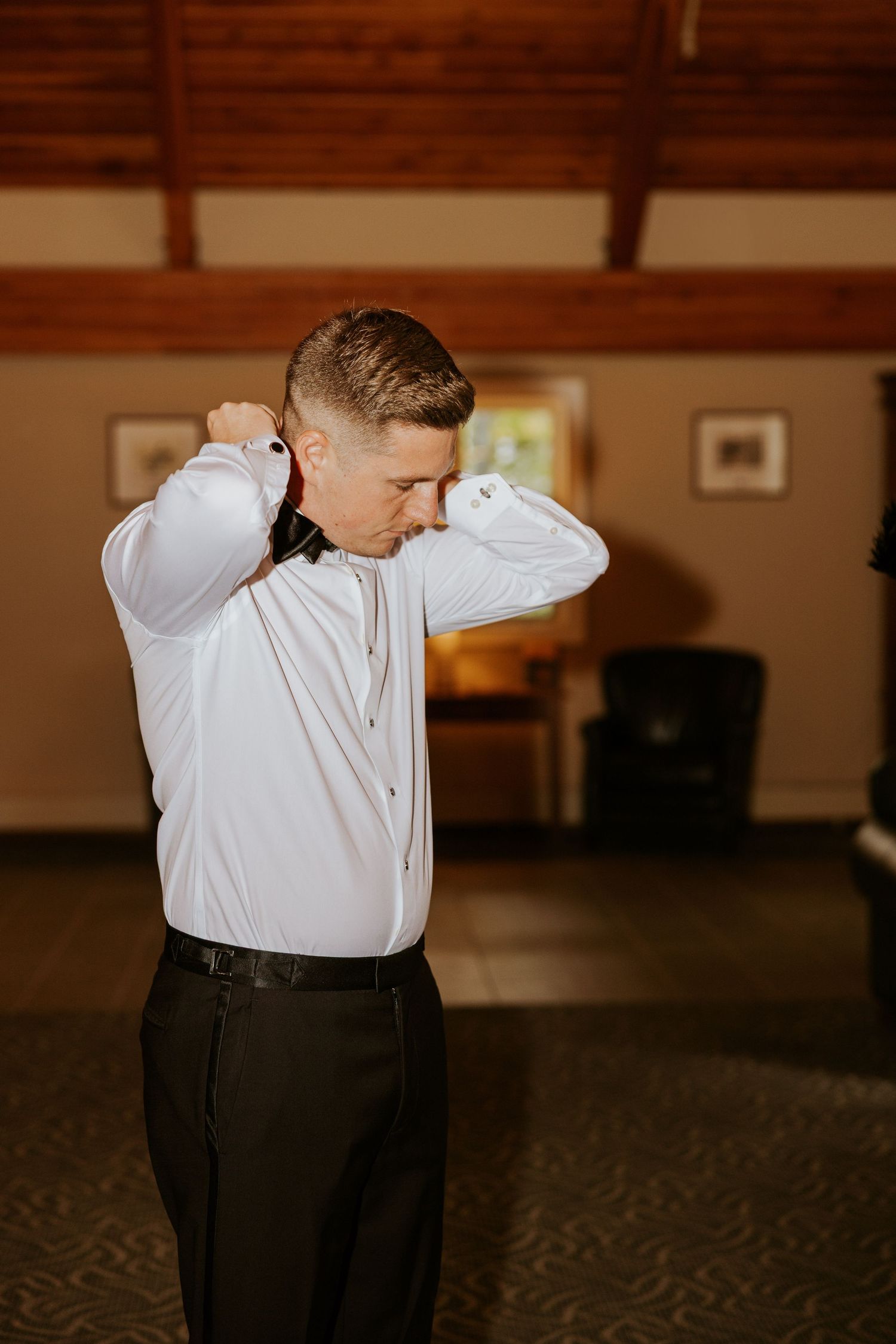 Groom in white shirt adjusts his black bow tie while getting ready for his wedding day.