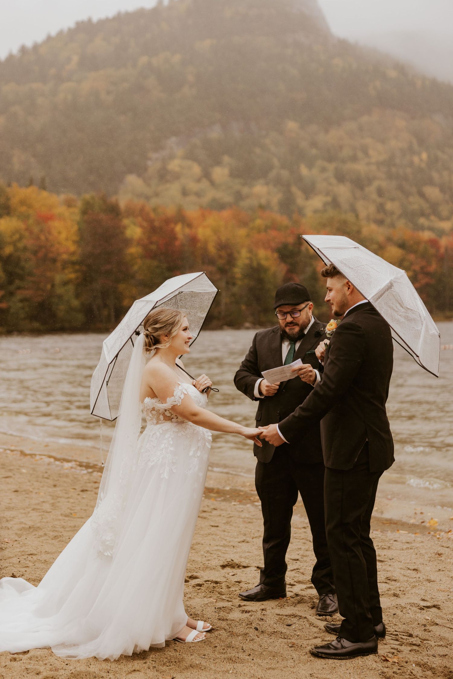 Couple exchanges vows on a foggy beach with colorful autumn mountains and an officiant behind them.