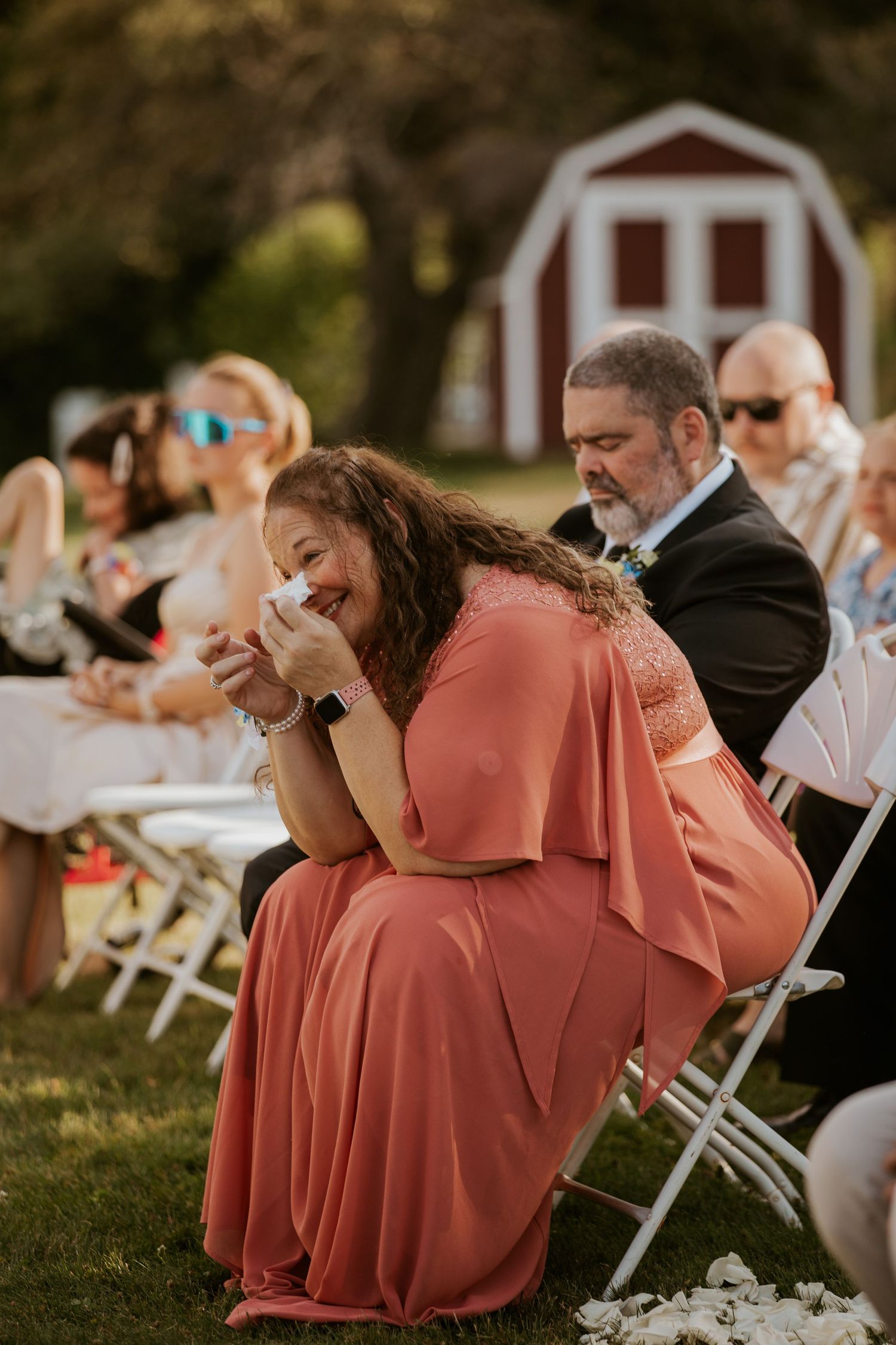 Emotional wedding guest in coral dress wiping tears of joy while seated at outdoor garden wedding ceremony.