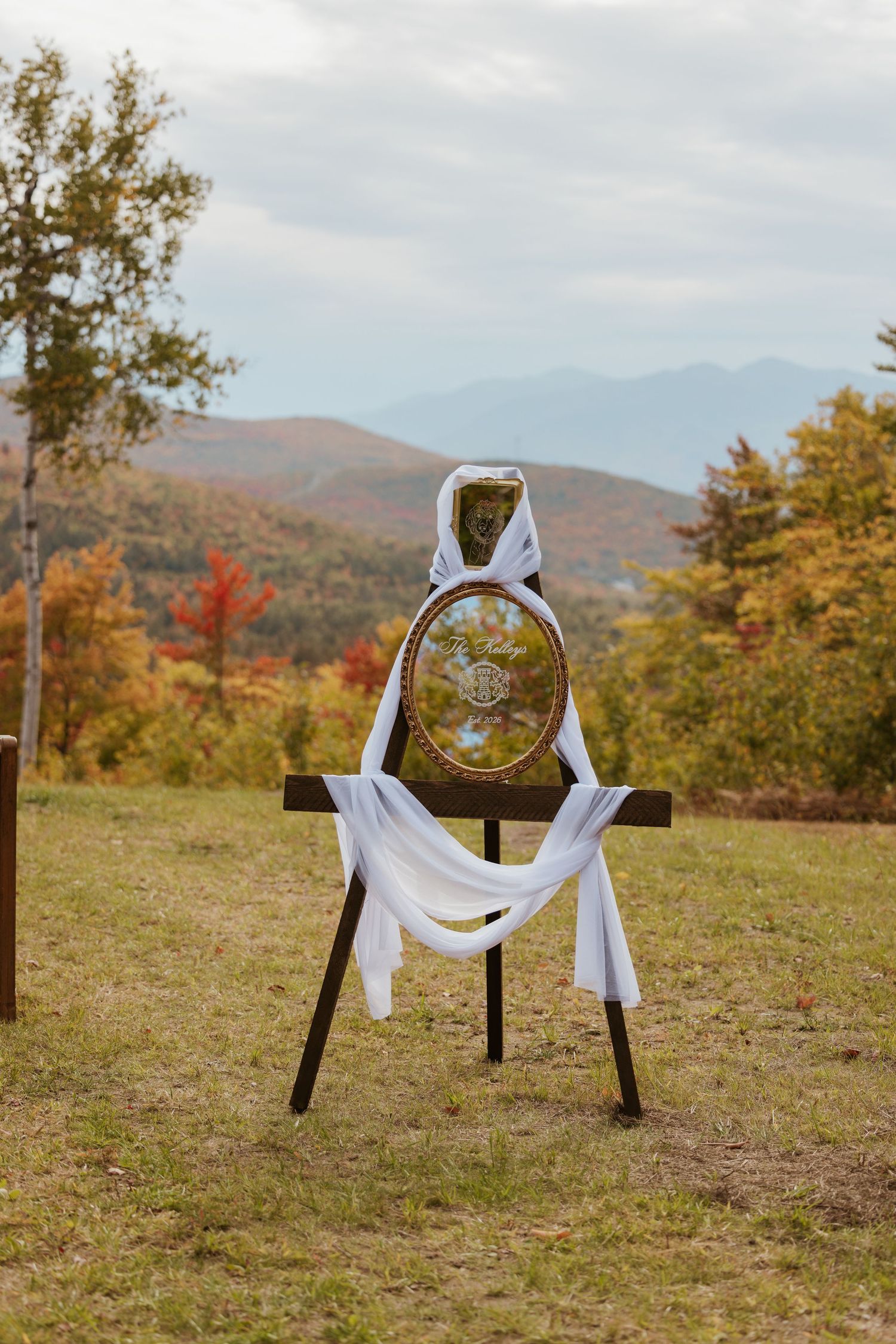 Rustic wedding sign draped in white fabric on wooden easel with autumn mountain landscape backdrop.