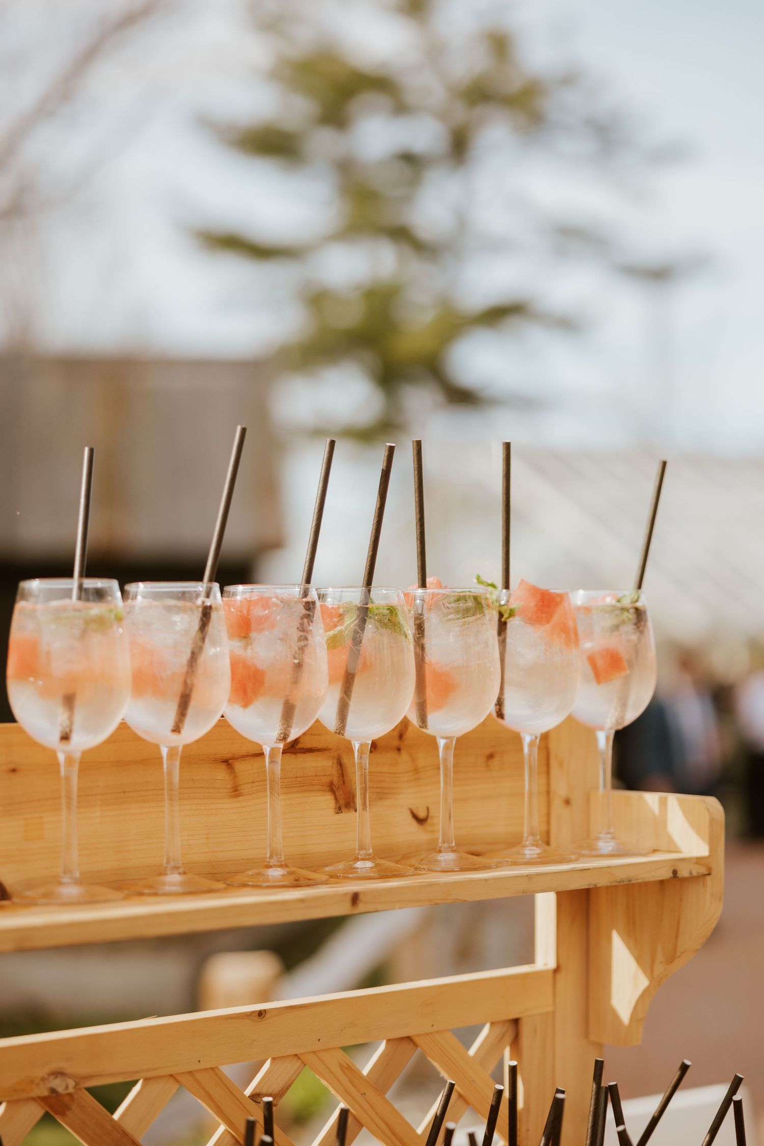 Row of elegant wine glasses with pink cocktails, ice, mint, and metal straws on a wooden bar cart outdoors.