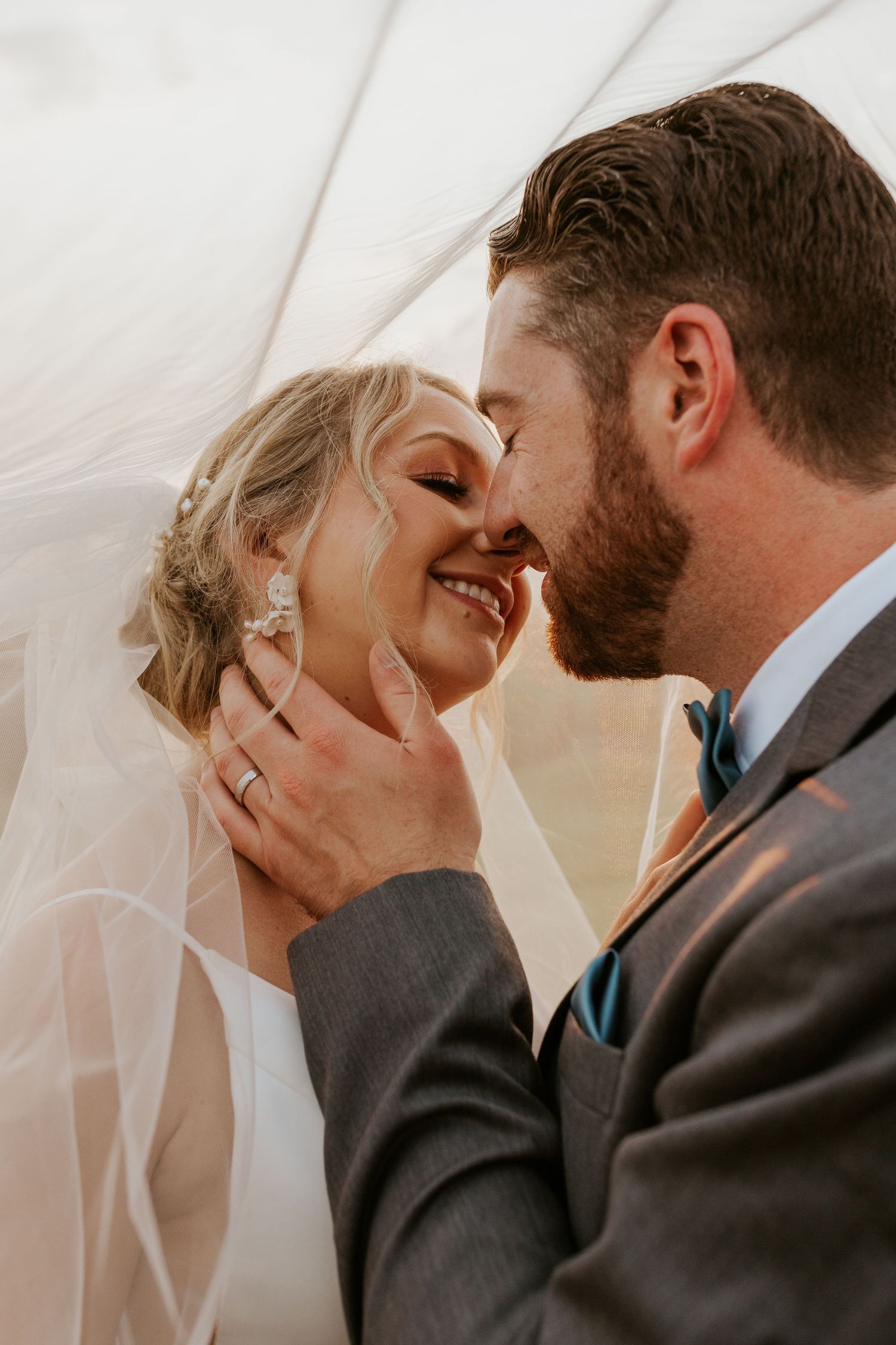 Groom tenderly holds brides face under flowing veil as they smile together closely.
