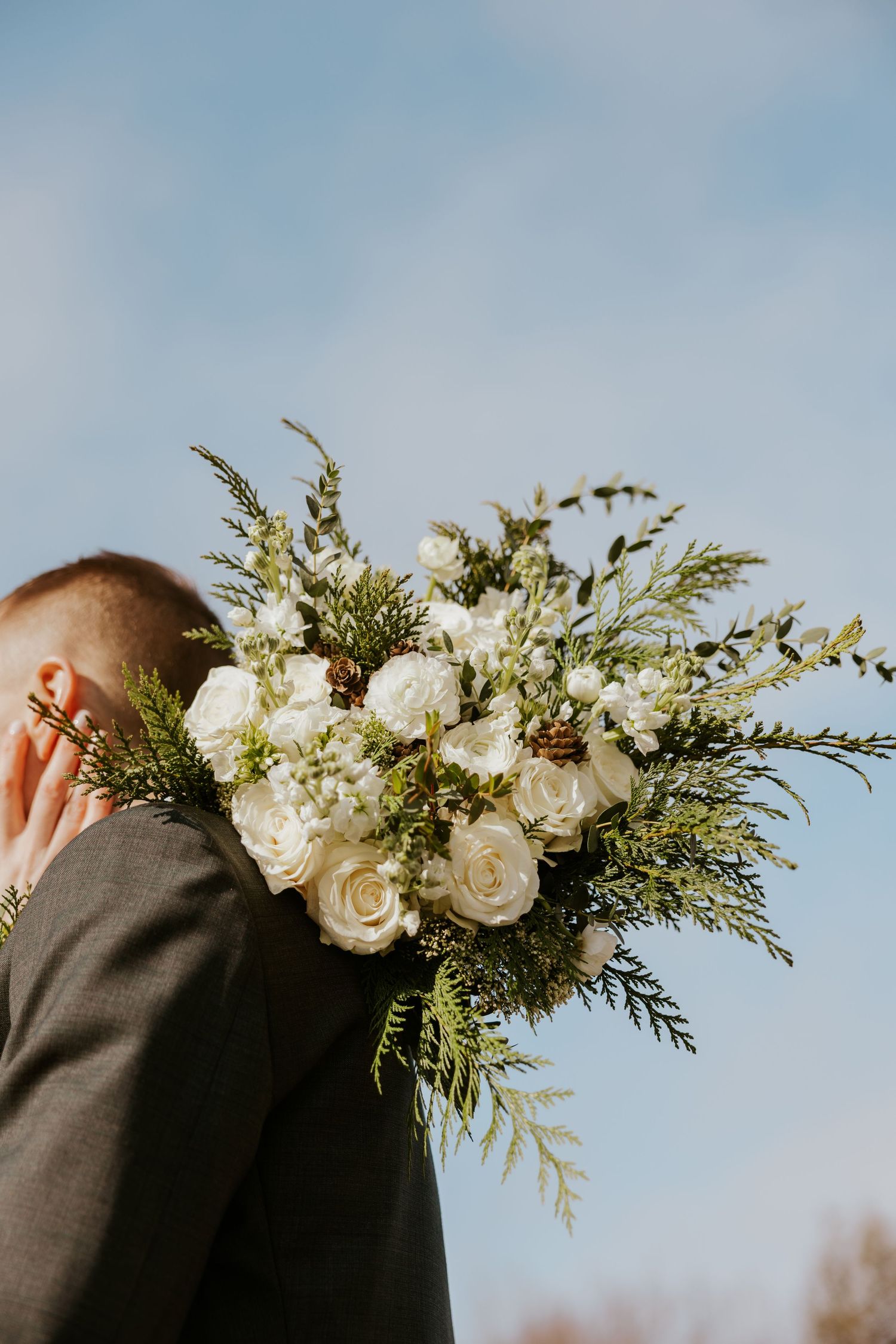 White roses and greenery boutonnieres displayed on dark suit jackets against a blue sky background.
