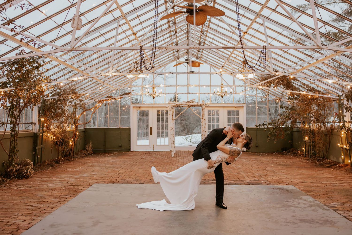 Groom dipping bride in romantic dance inside a warmly lit glass greenhouse venue.