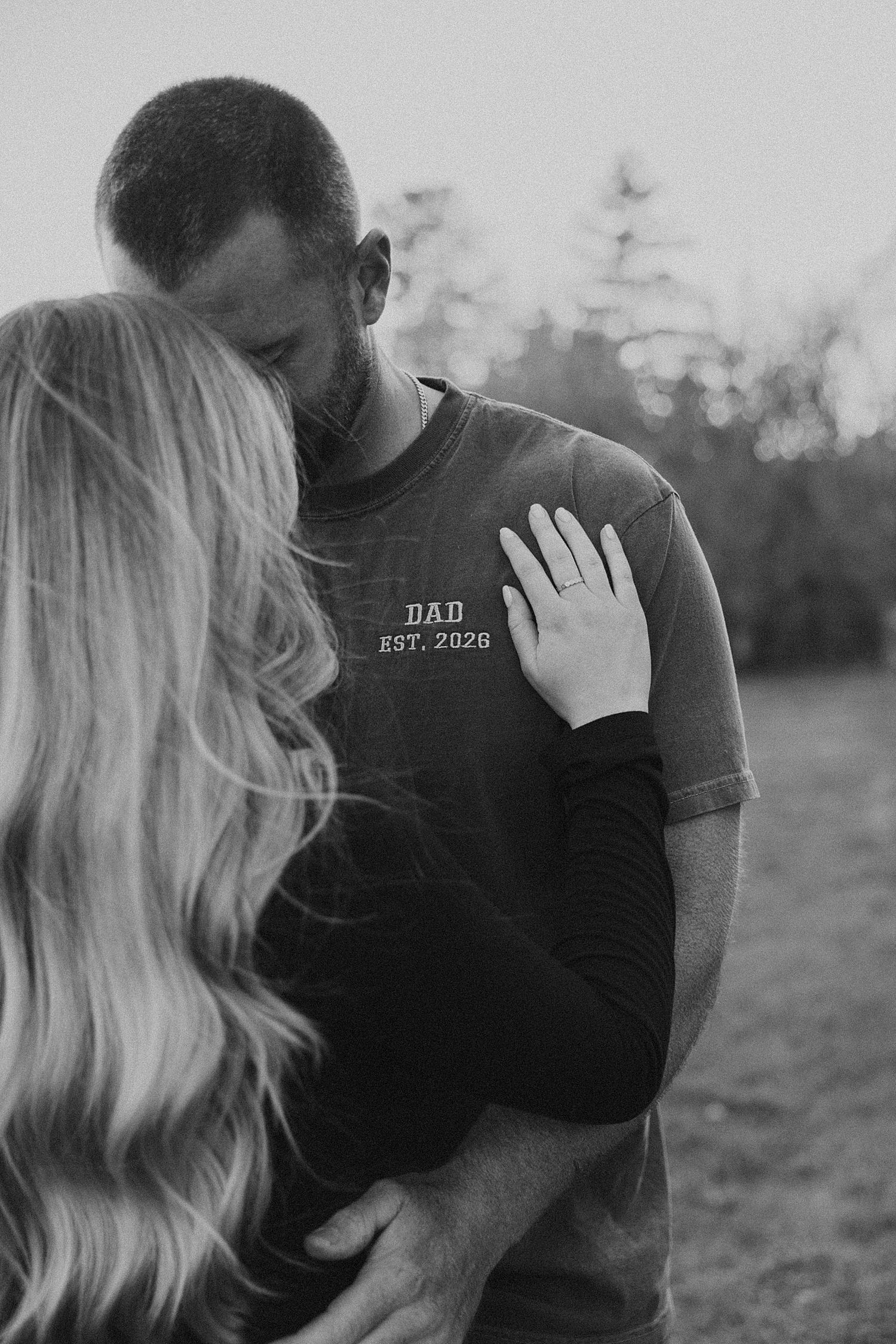 Black-and-white photo of a New Hampshire couple embracing, man wearing a DAD EST. 2026 shirt, woman's hand resting on his chest.