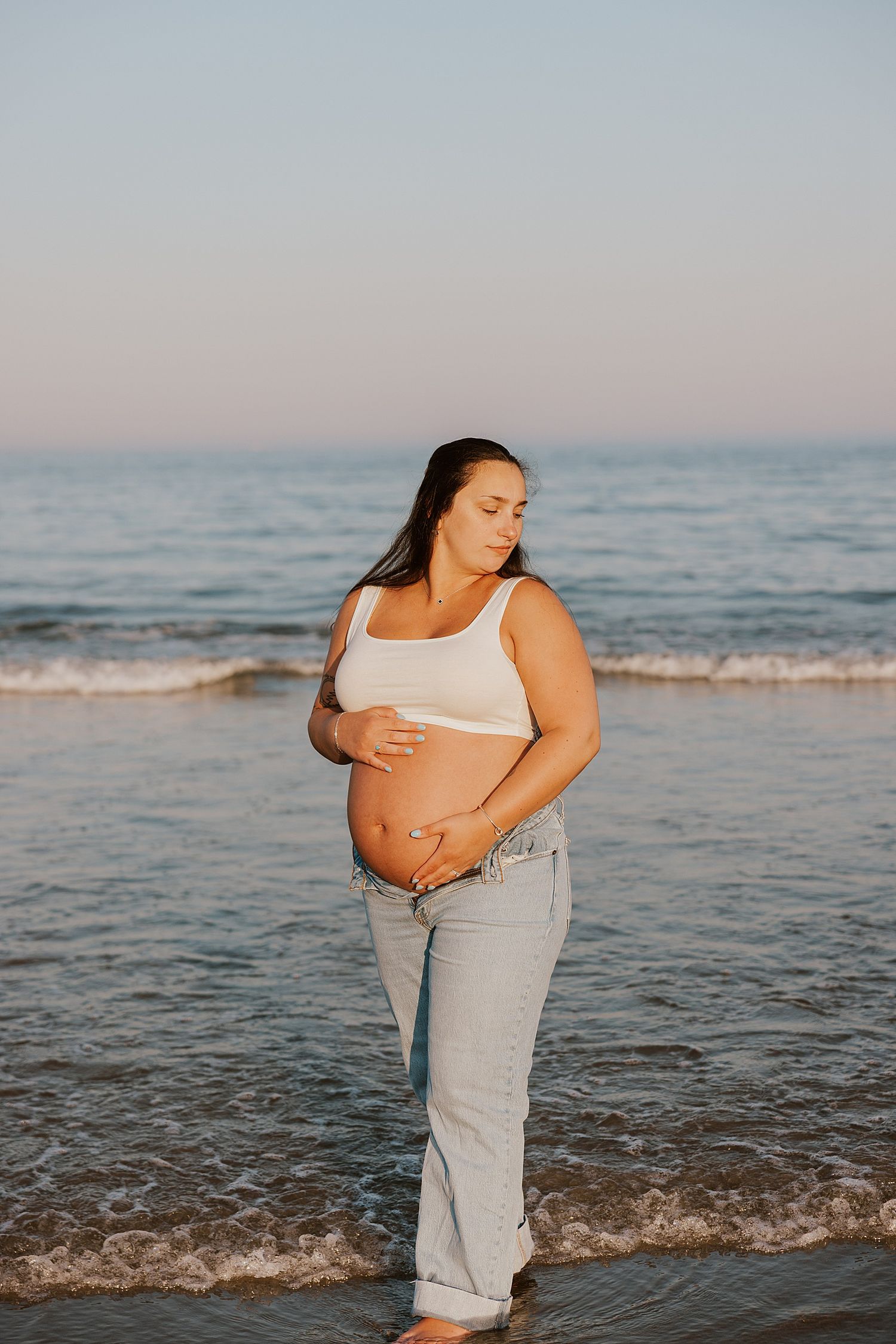 Pregnant woman in white crop top and jeans cradling her baby bump while standing in ocean waves at sunset in Rye, New Hampshire