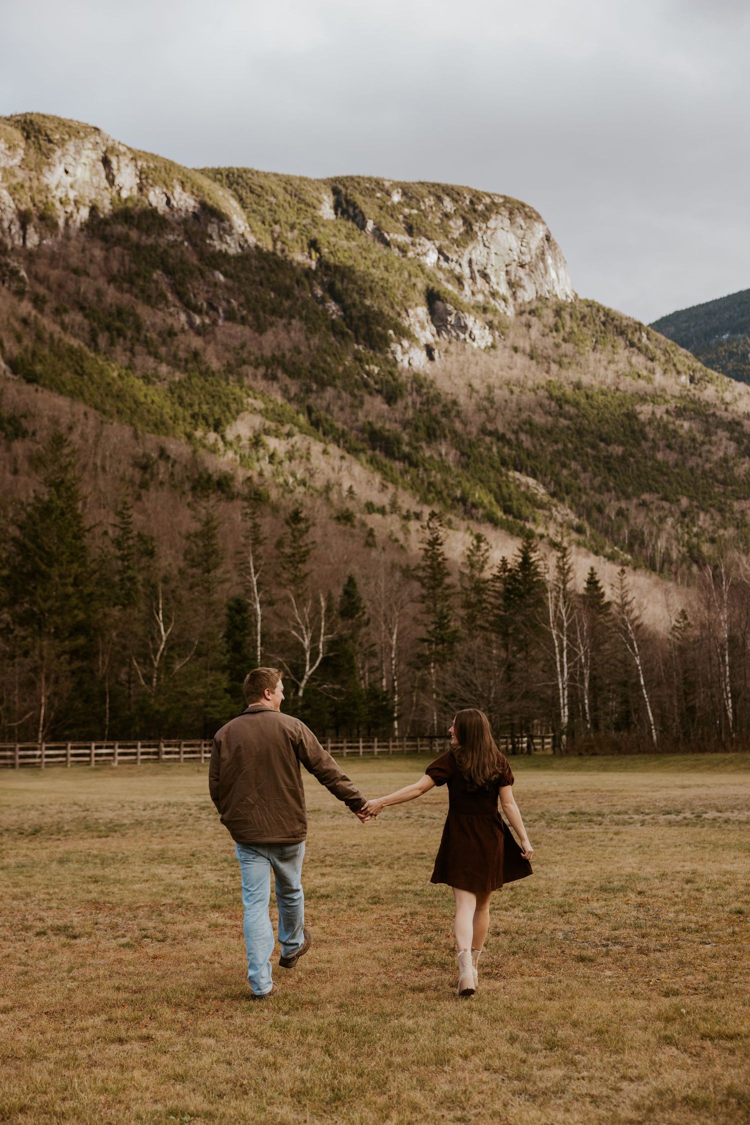 Couple walking hand in hand across a grassy field toward a stunning mountain backdrop with pine trees and a fence.