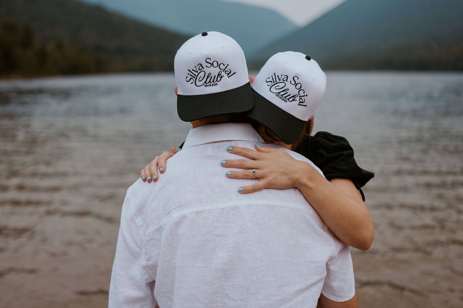Couple embracing from behind wearing matching white snapback hats near a mountain lake.