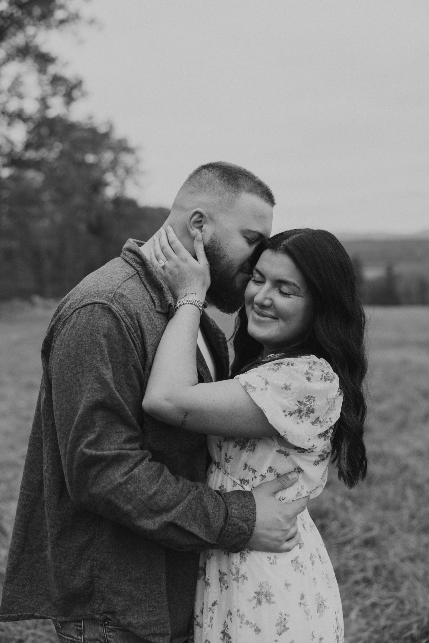 Black and white photo of man kissing womans cheek as she smiles joyfully in an outdoor fall setting.