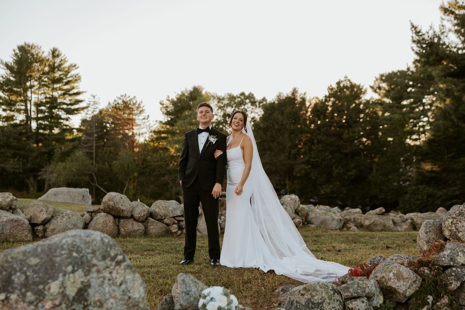 Bride and groom pose together among stone walls with pine trees at dusk.