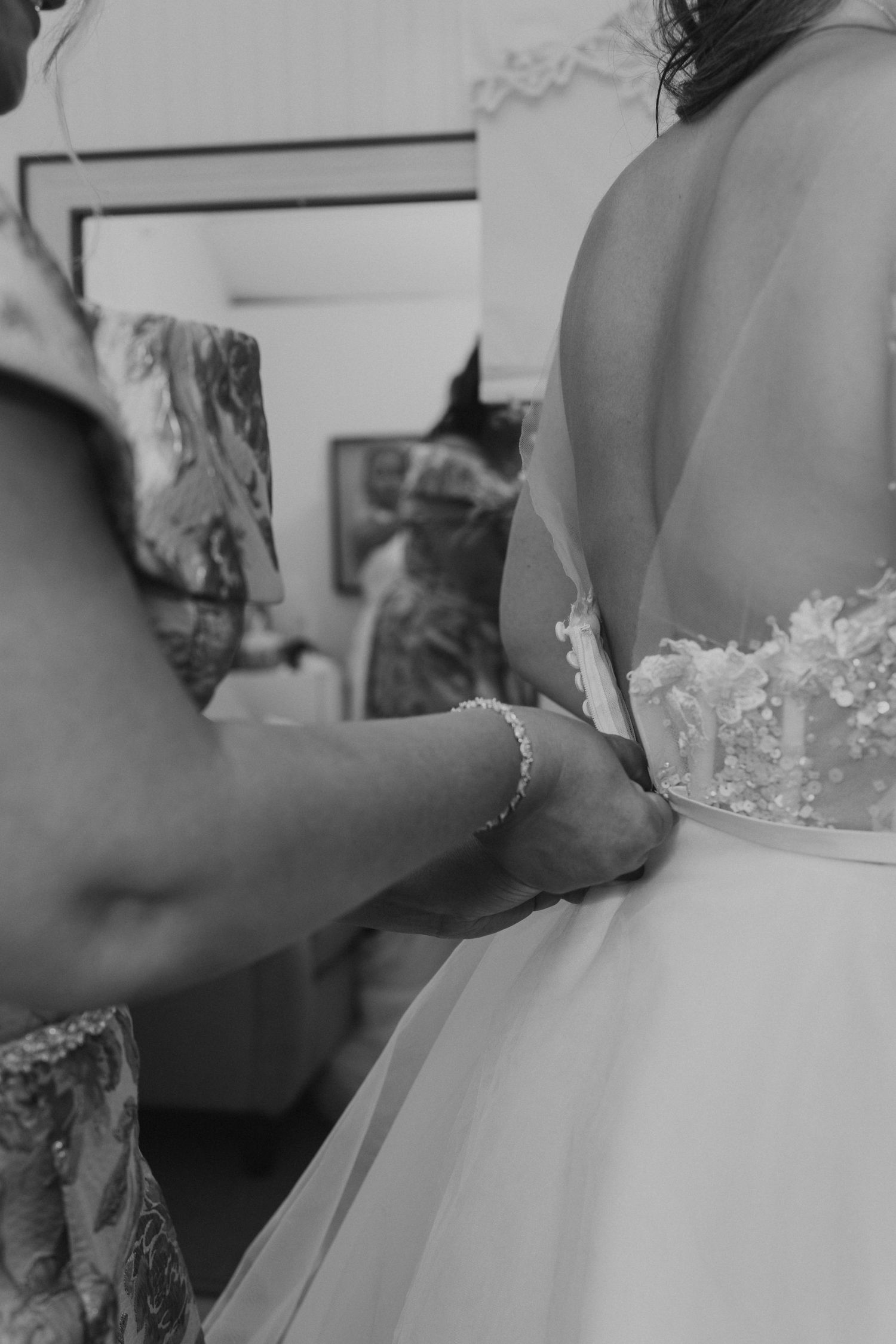 Black and white photo of someone fastening the back of a lace wedding gown on the bride.