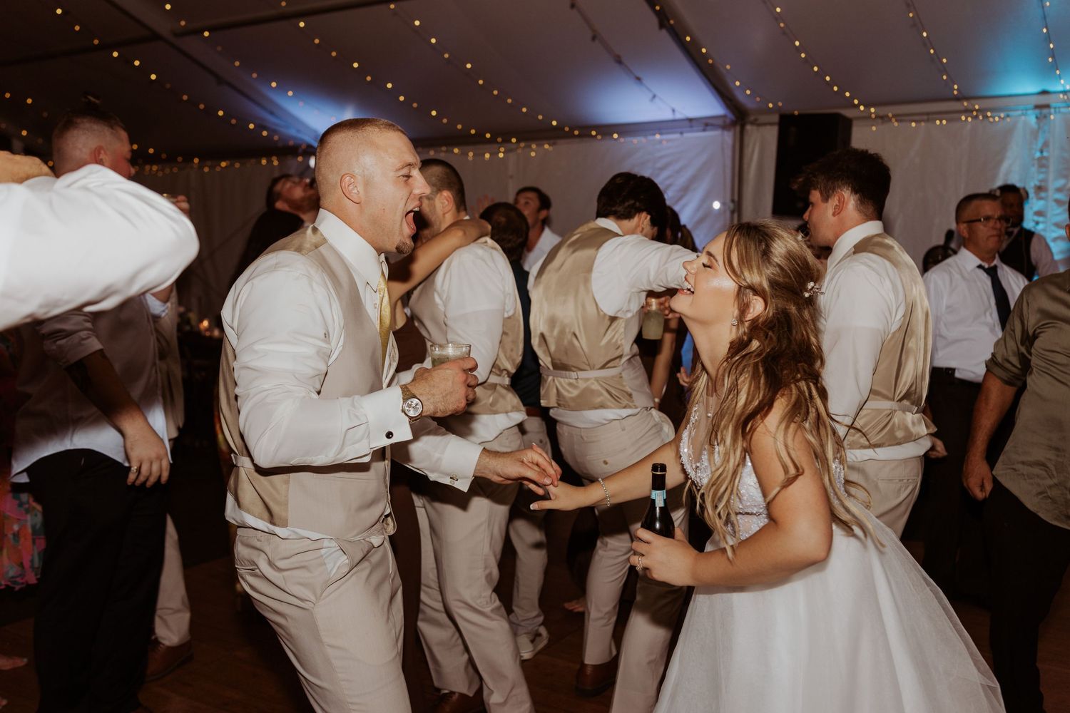 Bride and groom dance joyfully with wedding party guests under string lights at lively reception.