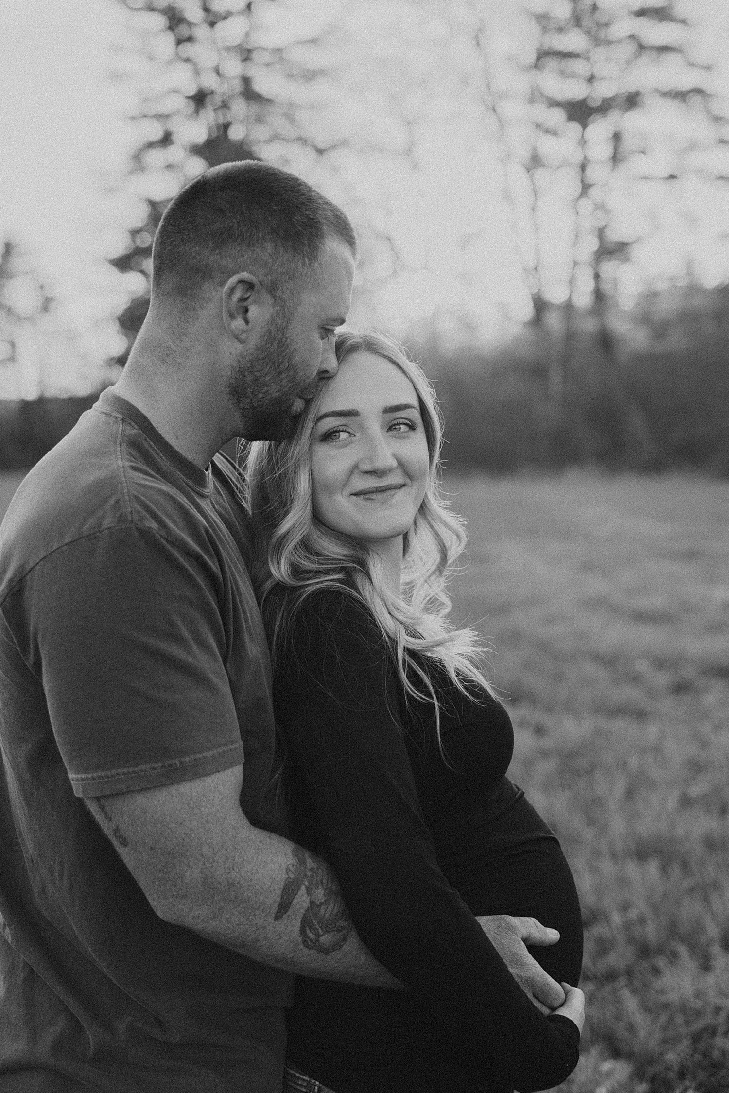 Black and white maternity photo of a New Hampshire couple embracing outdoors, man kissing woman's head while she smiles at camera.