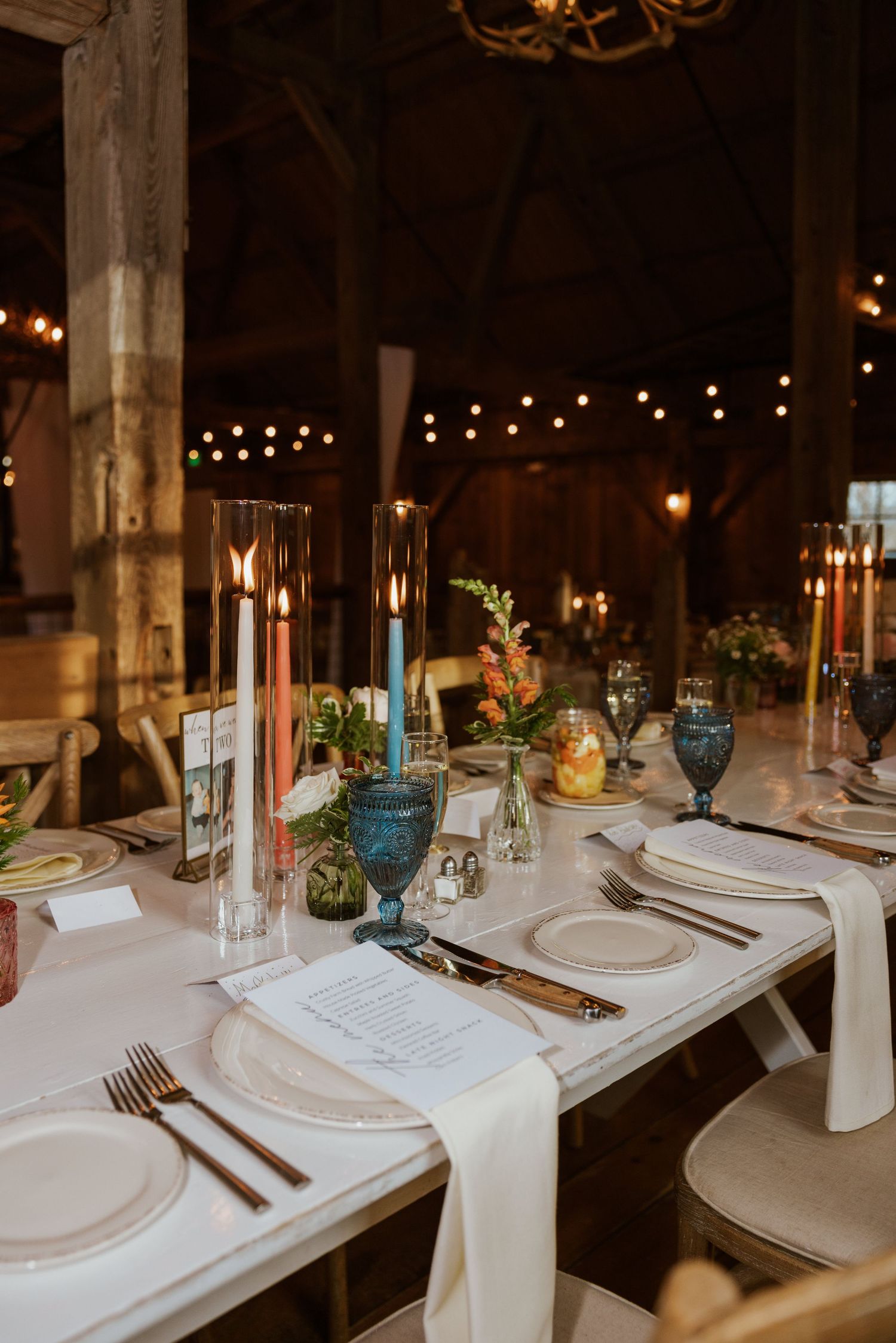Elegant barn wedding reception table with colorful taper candles, blue glassware, and string lights.