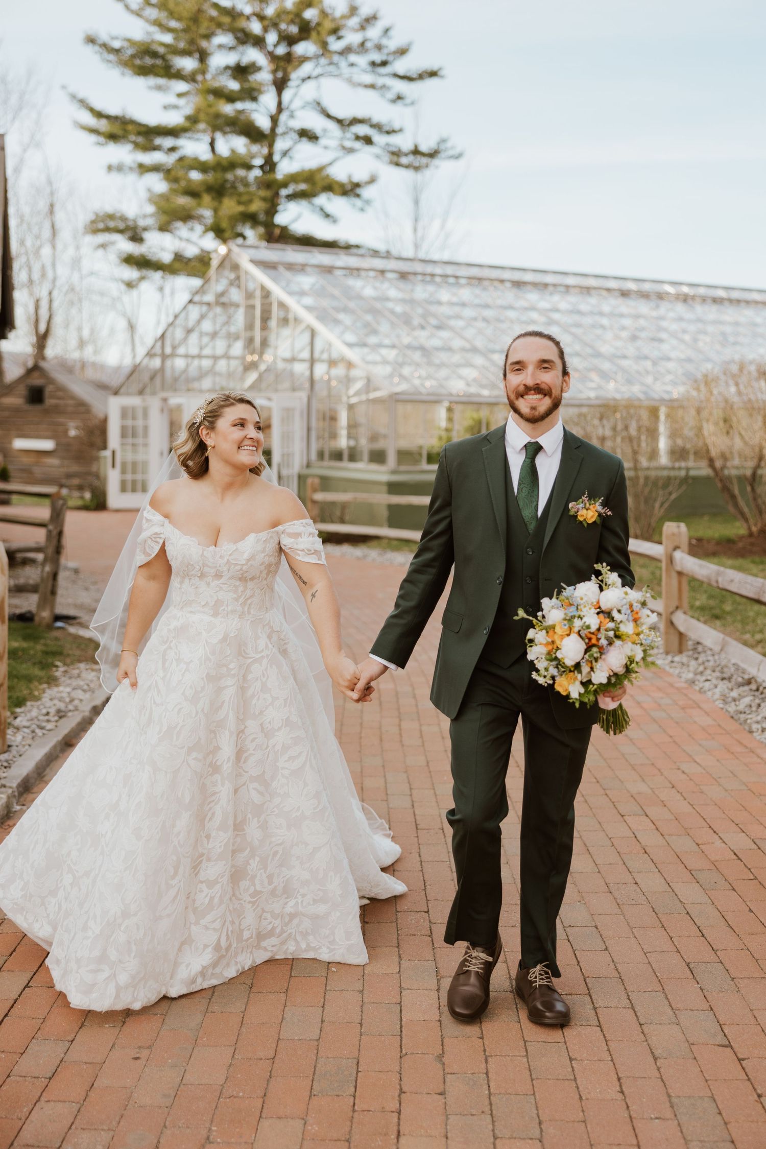 Bride in off-shoulder lace gown and groom in green suit hold hands at wedding venue.