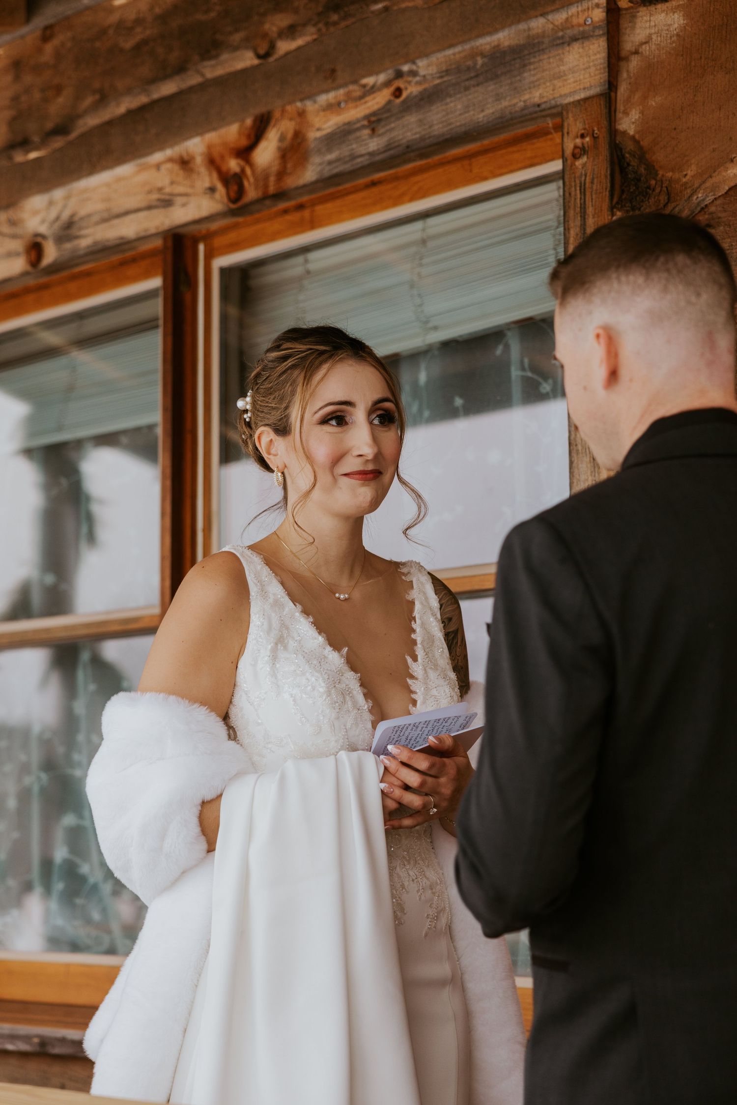 Bride in white lace gown and fur wrap reads vows to groom in black suit at rustic cabin wedding.