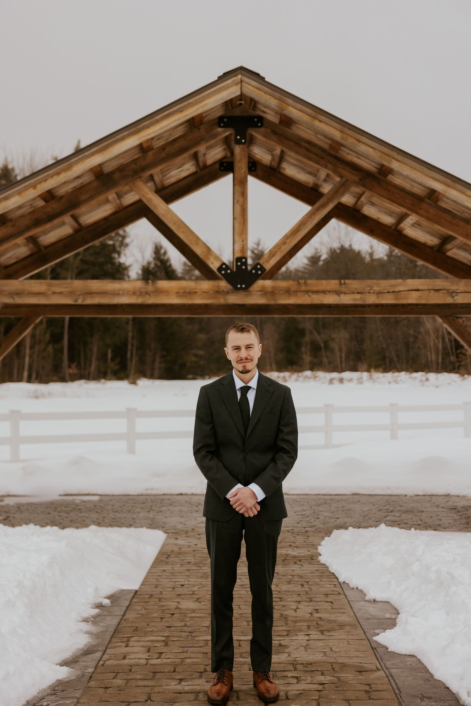A groom stands under a rustic wooden pavilion on a snowy winter day wearing a black suit.