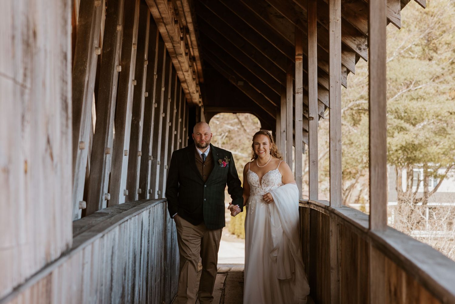 Wedding couple poses in rustic covered bridge with wooden beams and natural lighting.