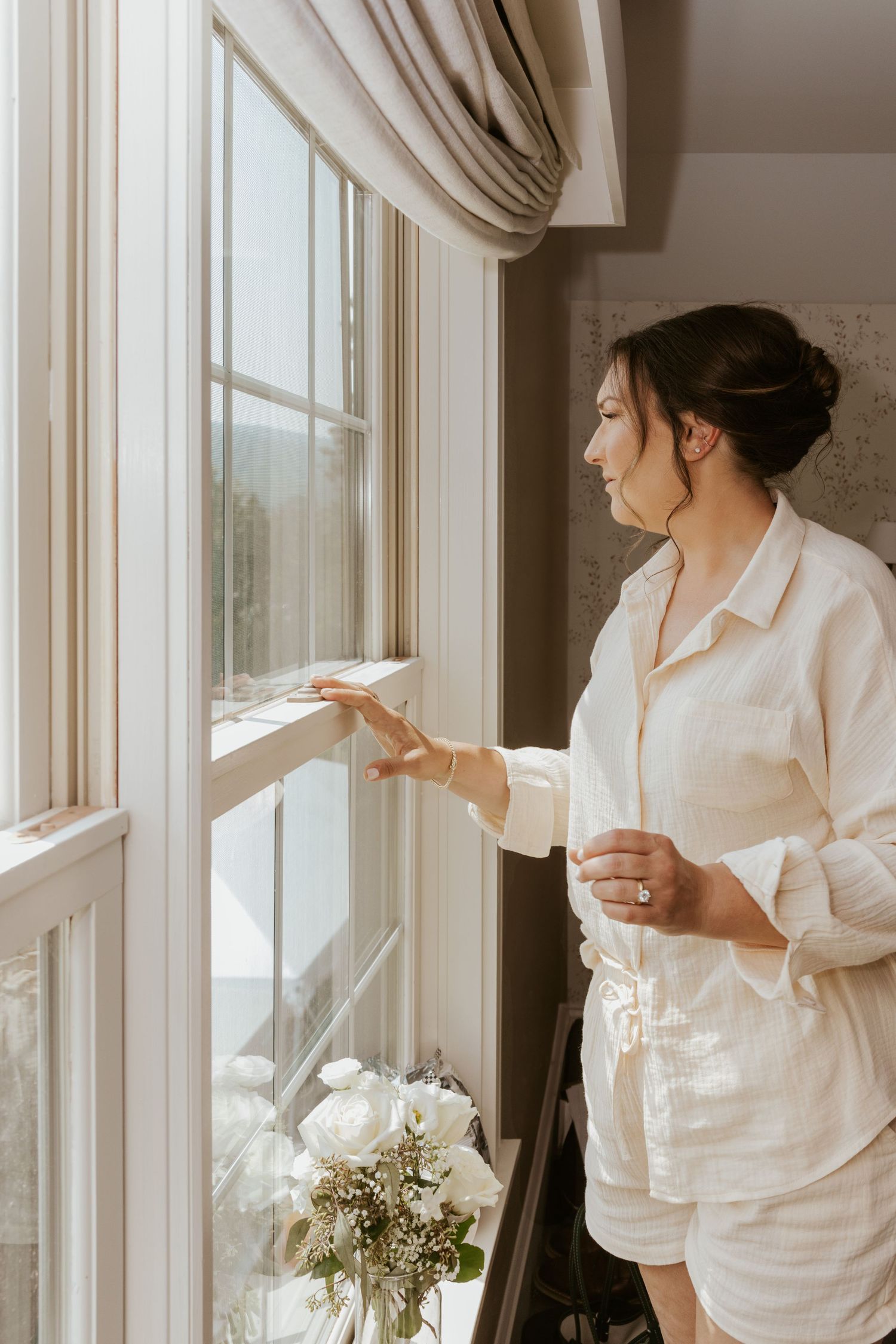 A bride in white pajamas smiles while looking out a bright window holding a coffee cup on her wedding morning.