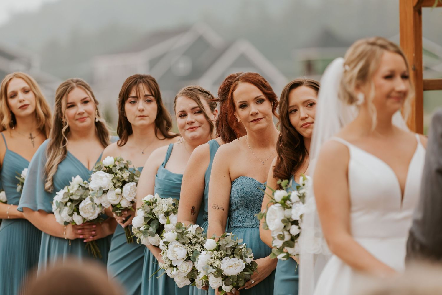 Bridesmaids in dusty blue gowns holding white floral bouquets stand beside bride during outdoor wedding ceremony.