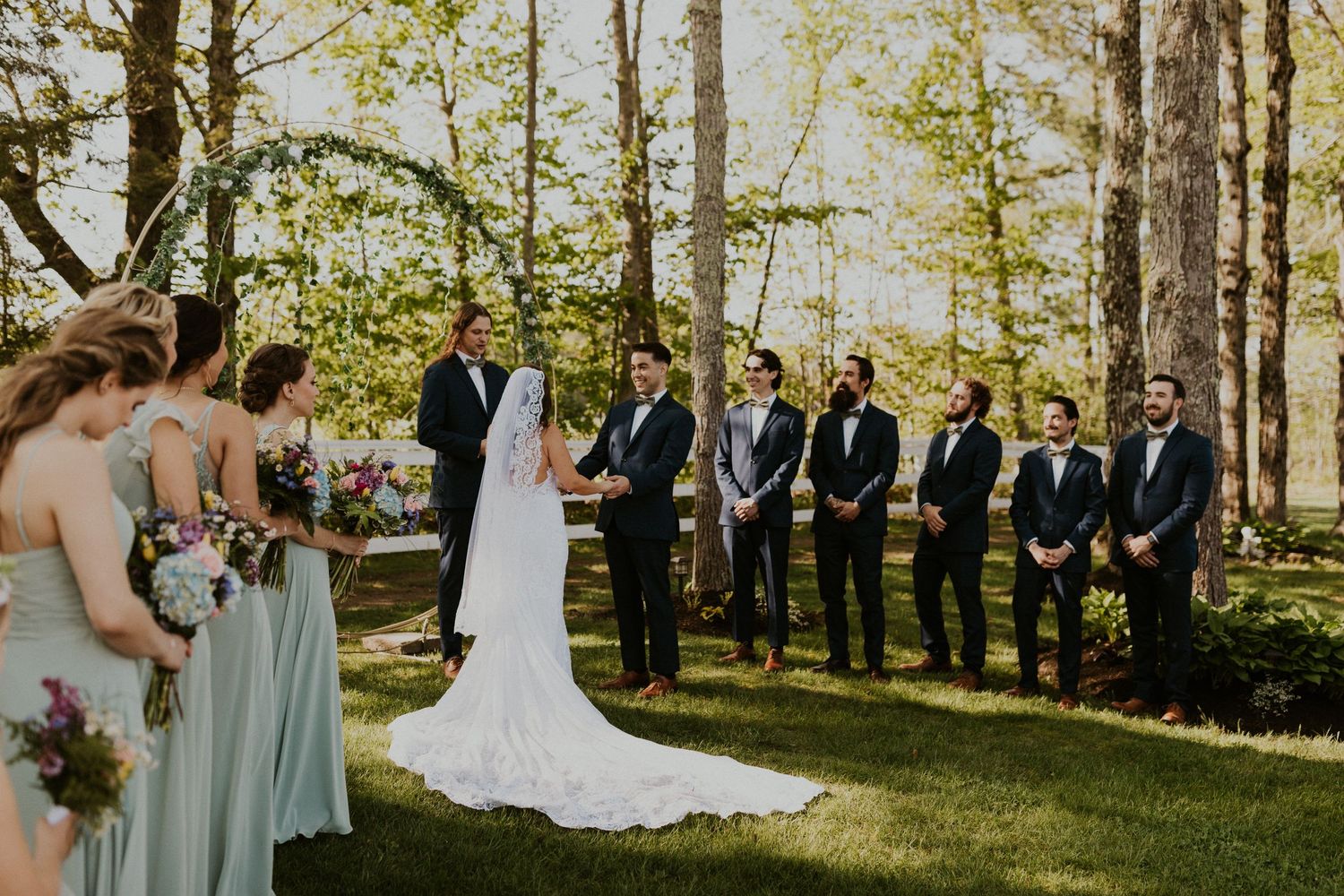 Chrissy and Mike holding hands as their wedding ceremony begins. It is a lovely spring day with the sun shining.