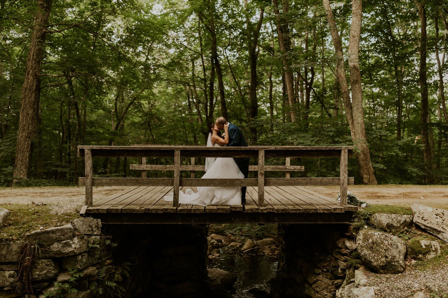 Stone wall and wooden bridge create rustic backdrop for outdoor wedding venue.