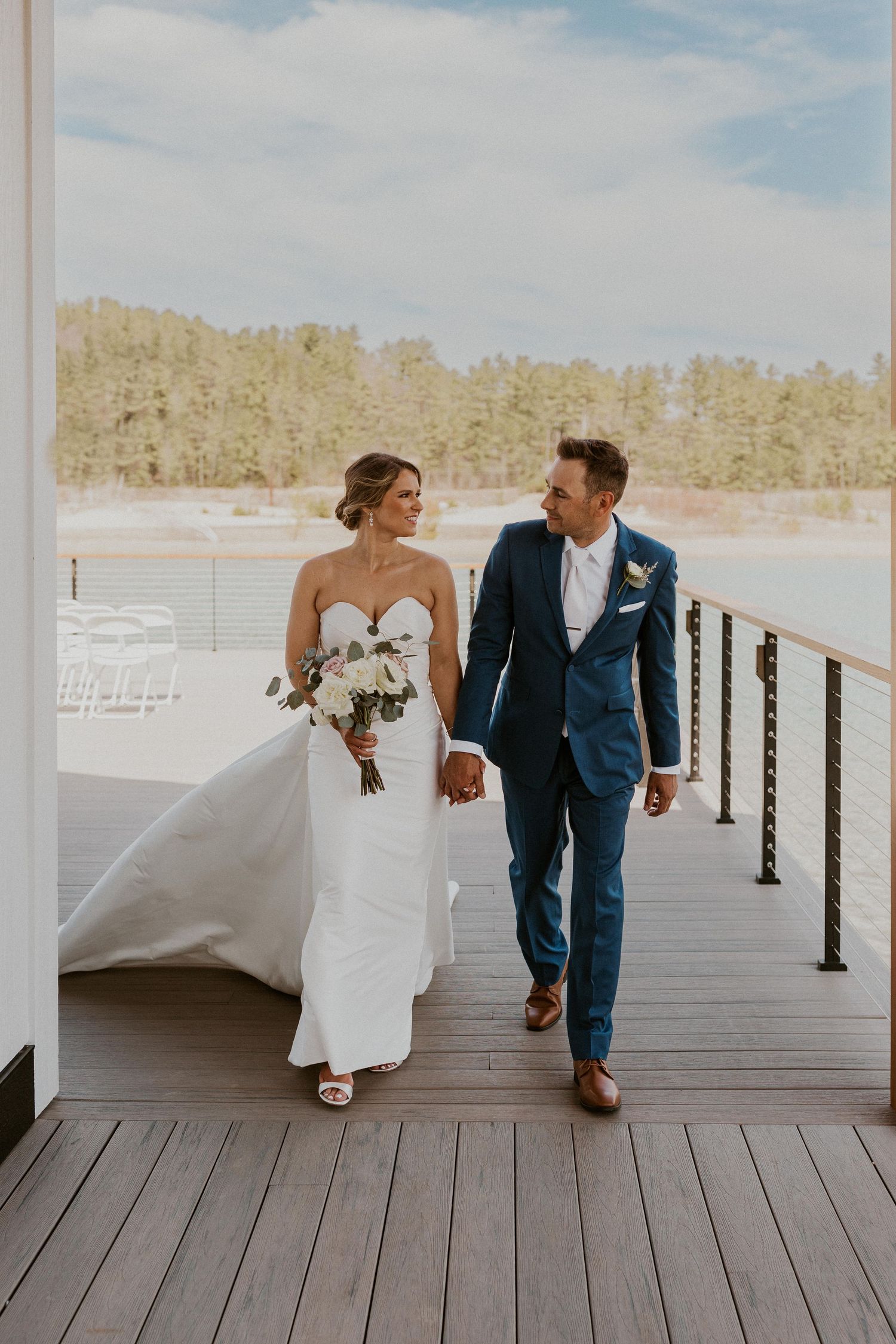Wedding couple walking on wooden dock by lakeside in formal attire.