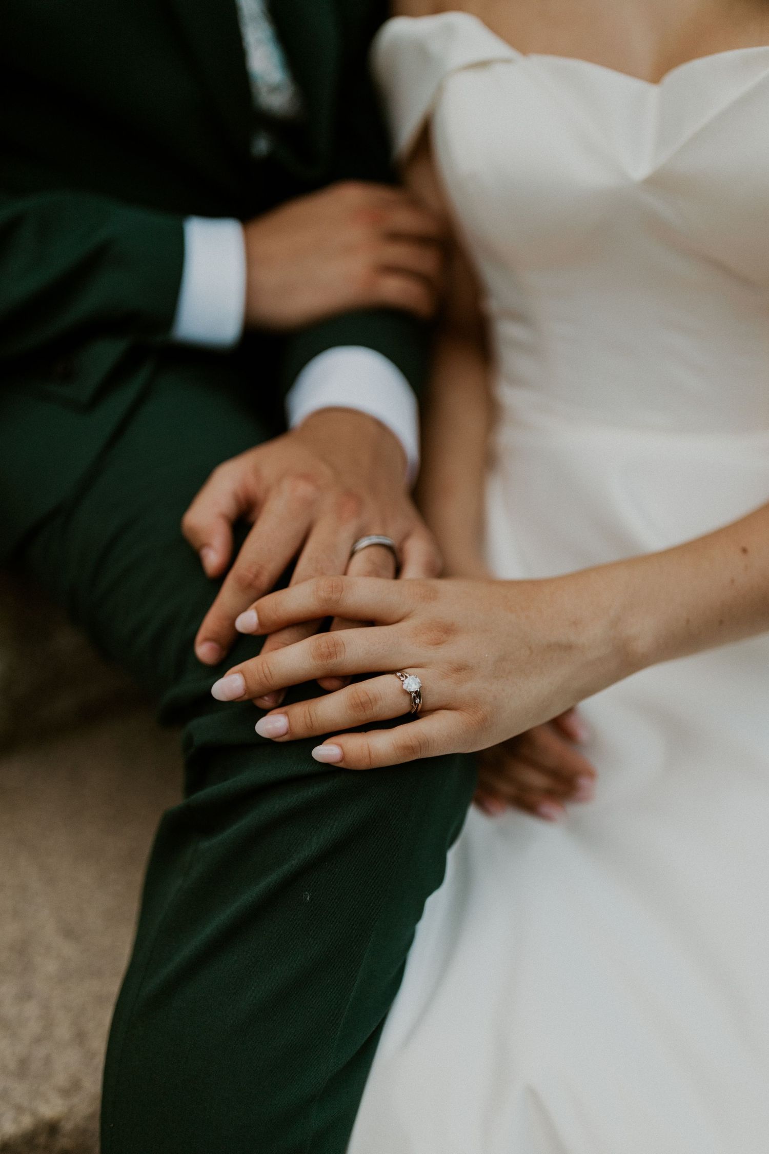 Close-up of wedding rings and hands against white dress and emerald green suit fabric.