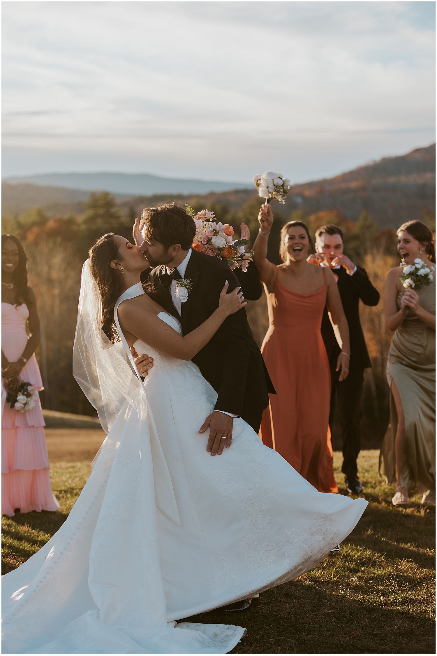 A wedding couple shares a romantic kiss at sunset on a hilltop while bridesmaids in colorful dresses look on in the background.