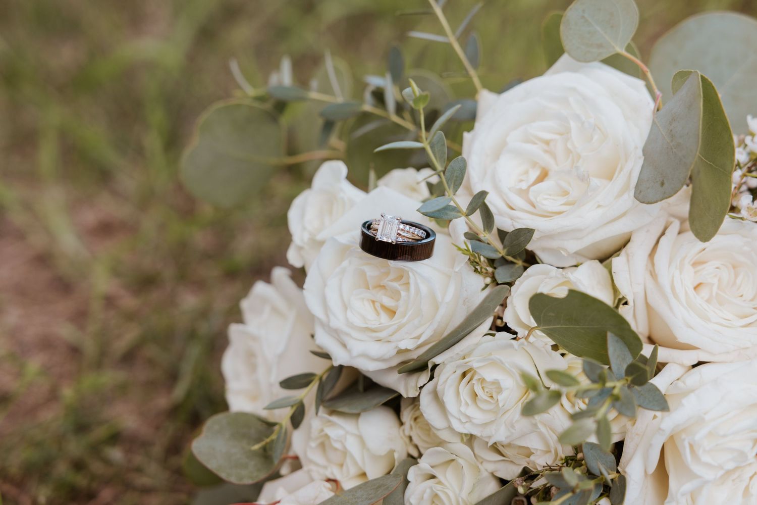 Wedding rings rest on a bouquet of white roses and eucalyptus greenery outdoors.